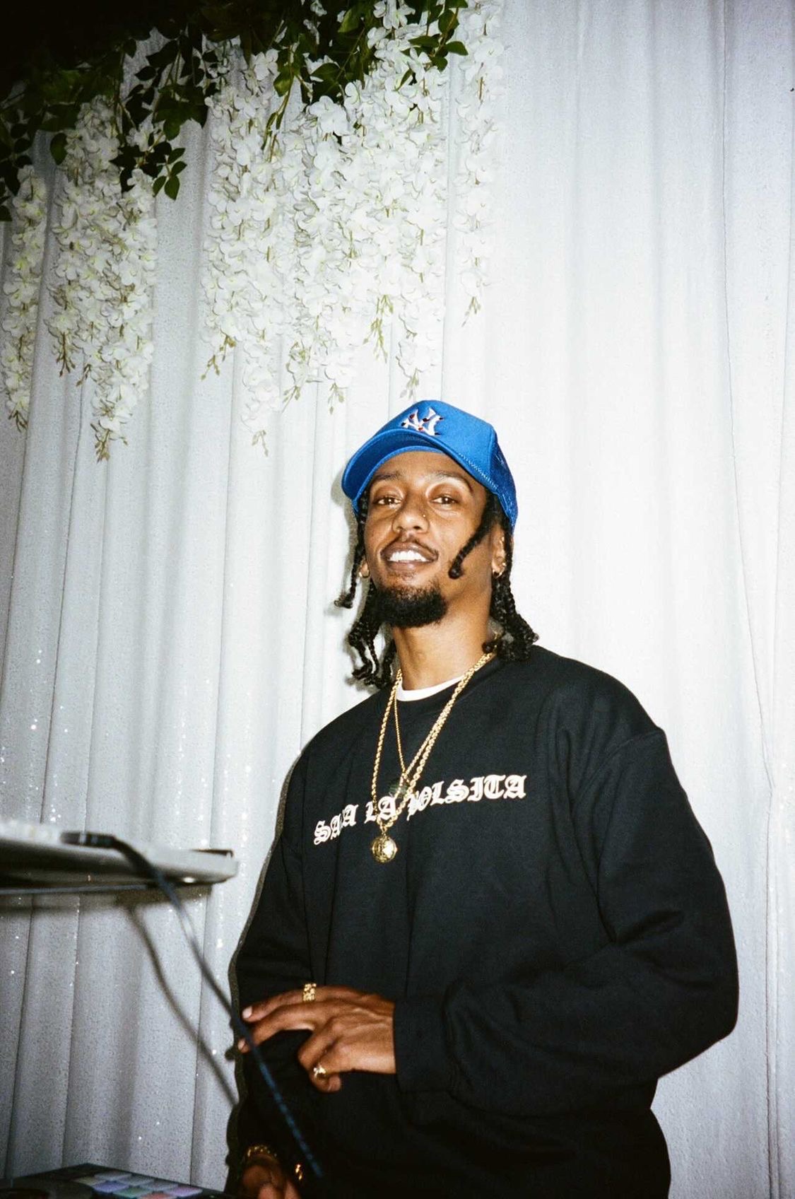 Man with a blue cap, gold chain, and black sweatshirt smiling in front of a white curtain with hanging flowers.