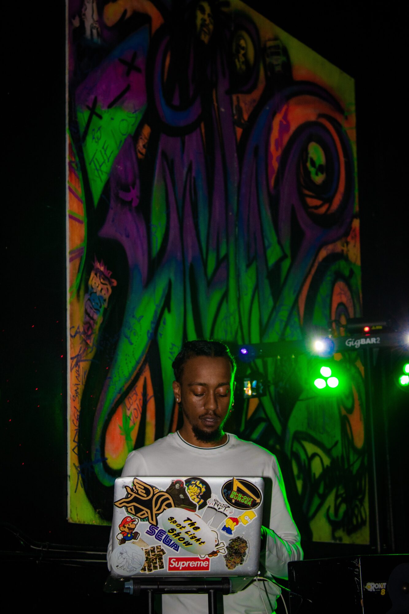 Person with a laptop decorated with stickers, standing in front of a colorful graffiti mural, illuminated by green stage lights.