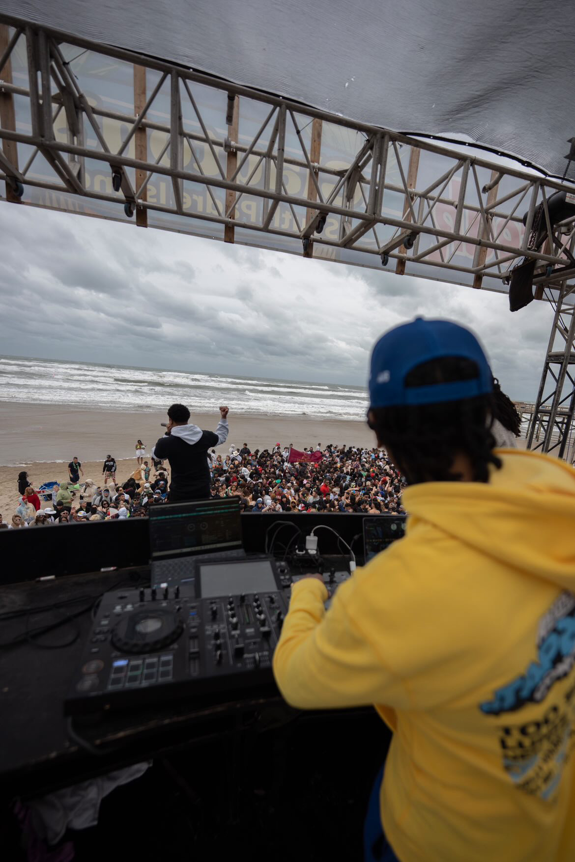 DJ in yellow hoodie and blue cap performs at outdoor concert with crowd and ocean in background.