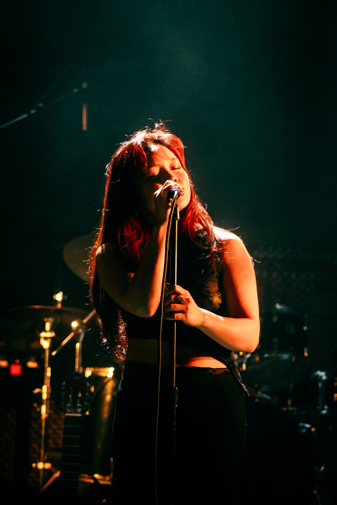 A woman singing into a microphone on stage with dark background and musical instruments, illuminated by stage lights.