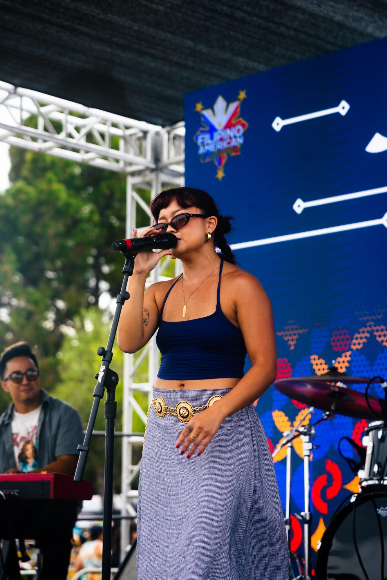 Woman singing into microphone on stage with a blue background and a person playing keyboard nearby.