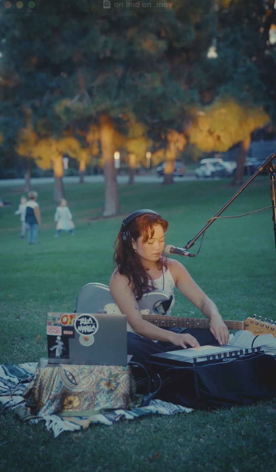 Girl playing keyboard outdoors with trees and people in background, sitting on a blanket, with a microphone nearby.
