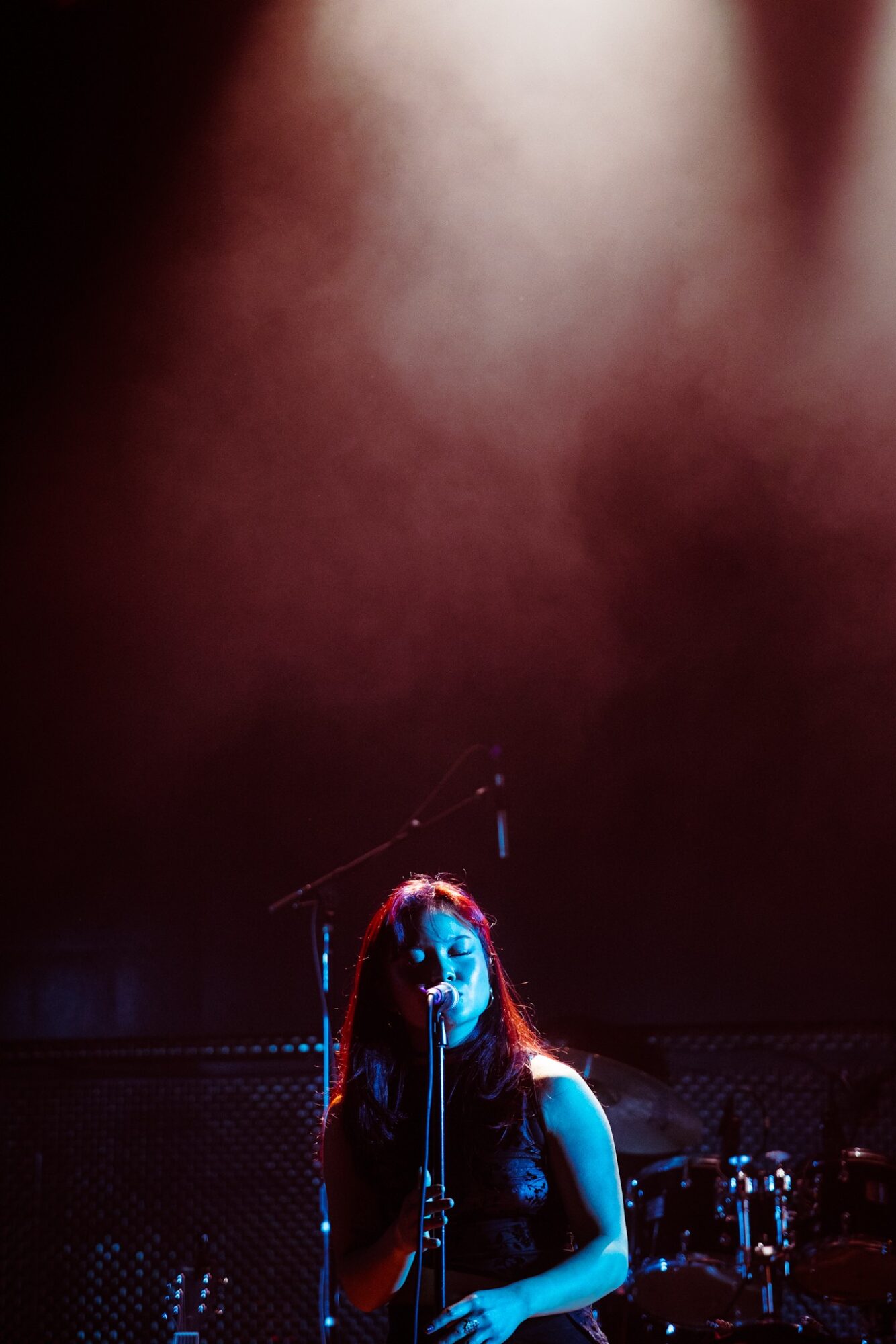 A person with long hair and glasses stands on stage with a microphone, illuminated by blue and red lights.