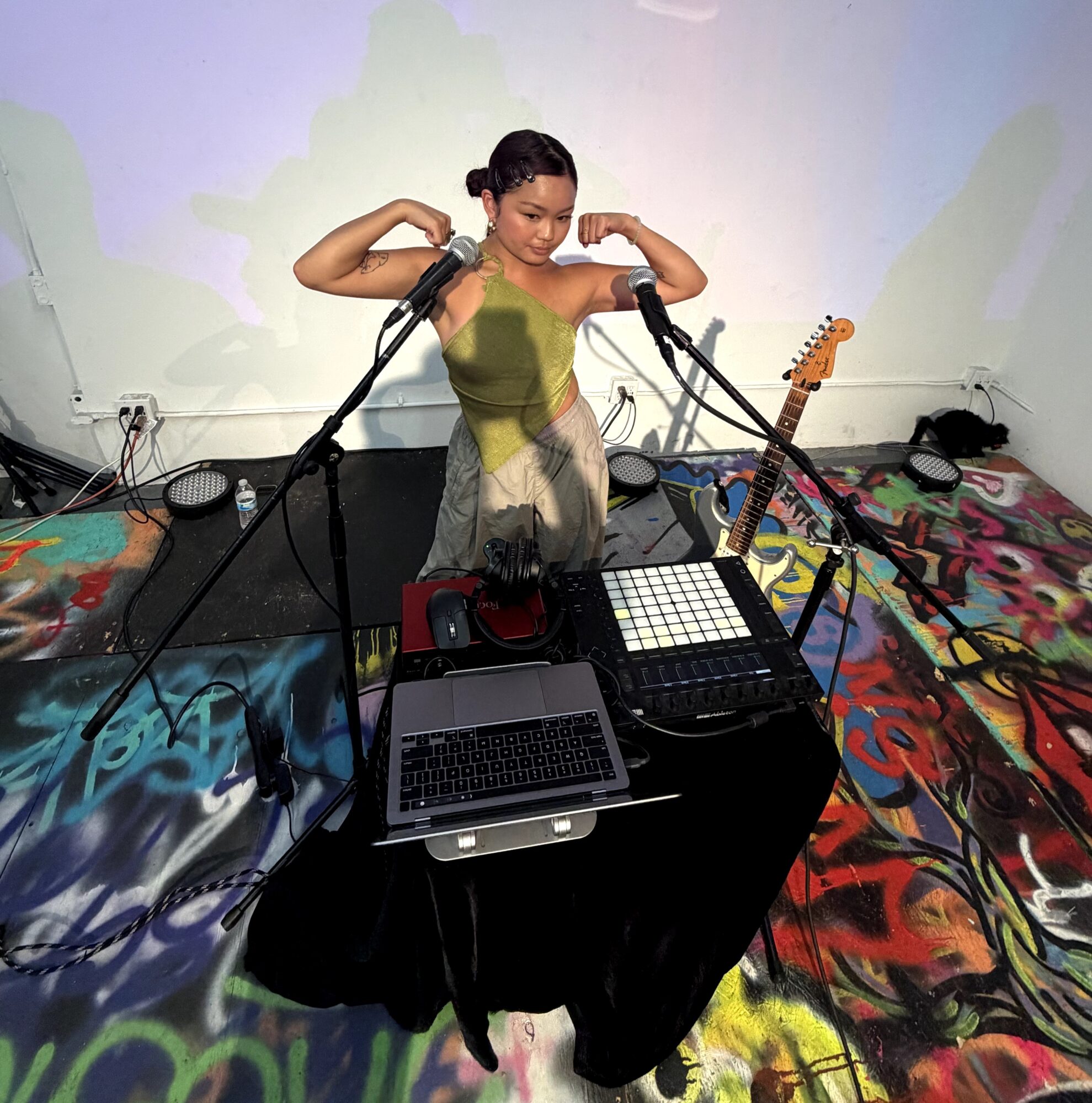Young girl with braided hair flexing her arms in front of a DJ setup with a laptop, mixer, and guitar, on a colorful floor.