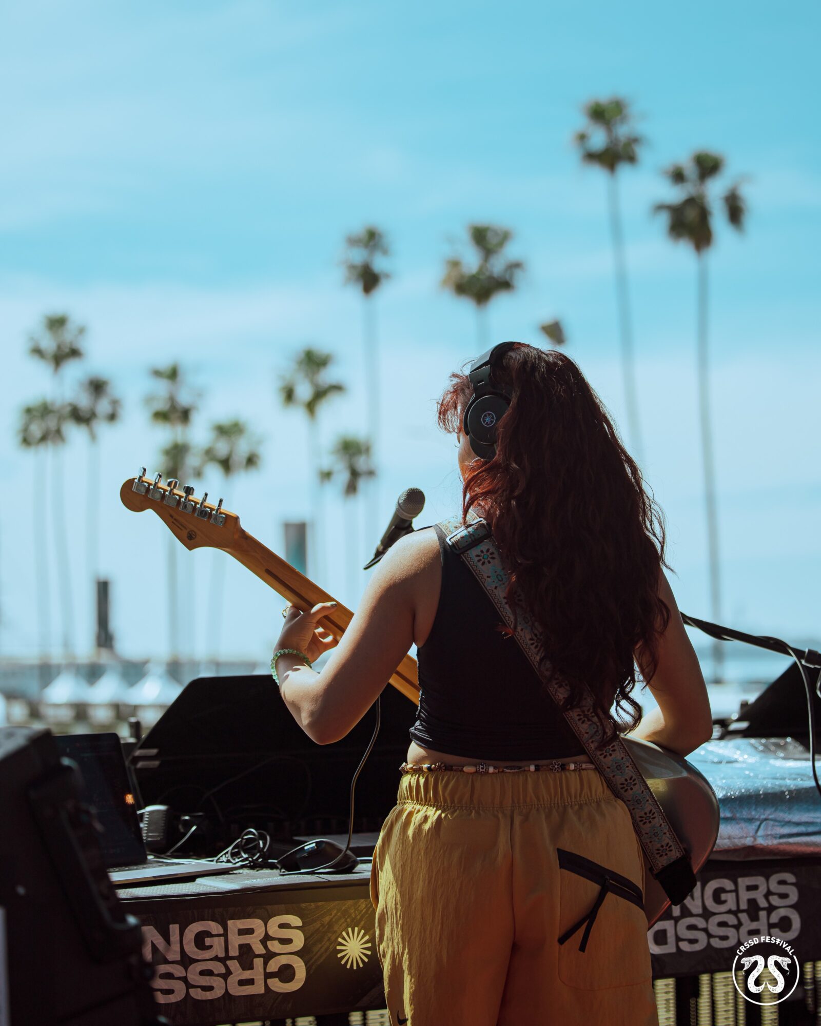 Person with long hair playing guitar outdoors with palm trees and blue sky in background.