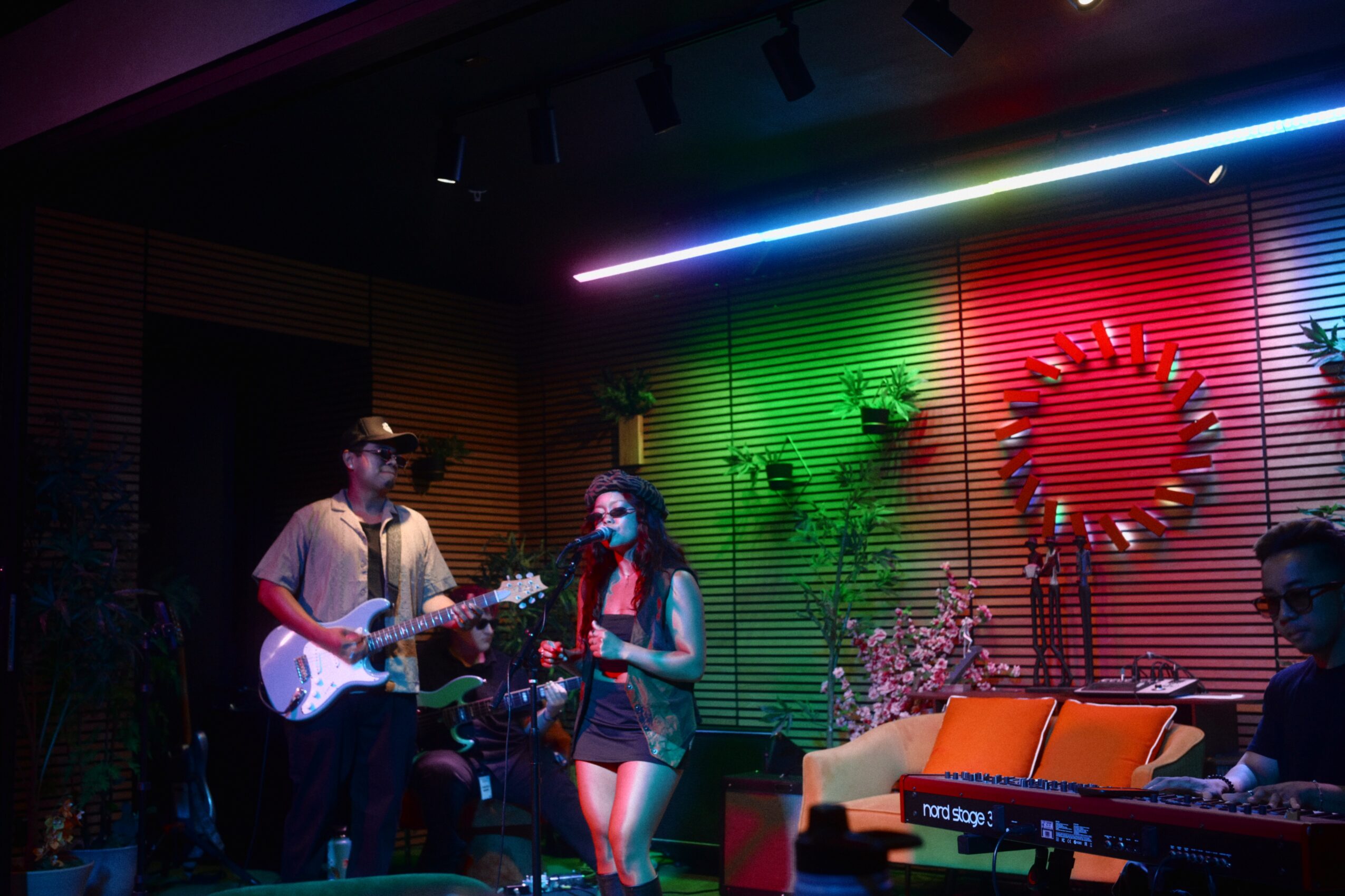 Musicians perform on stage with colorful lighting and a decorated backdrop, including a circular wreath and plants.