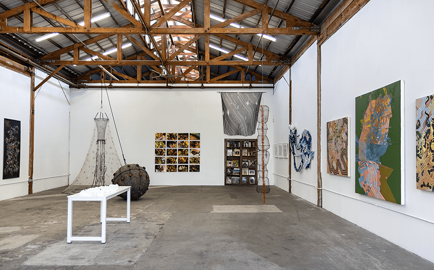Art gallery with white walls, wooden ceiling, and various paintings and sculptures displayed, including a wire sculpture and a table.