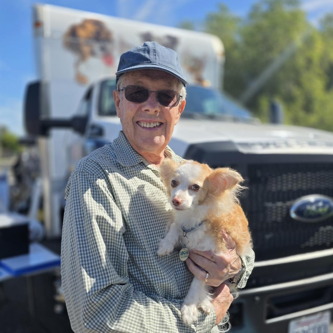 Man wearing sunglasses and a cap holding a small dog outdoors with a truck in the background.
