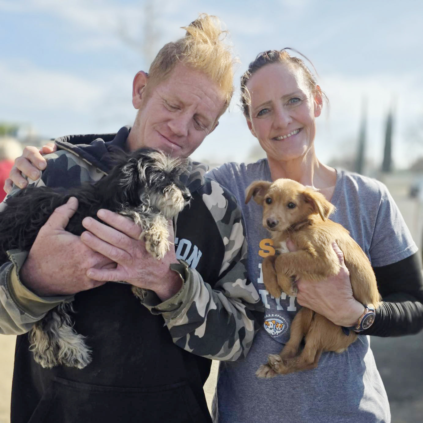 Two people holding puppies outdoors, one with a black and white puppy, the other with a brown puppy, smiling.