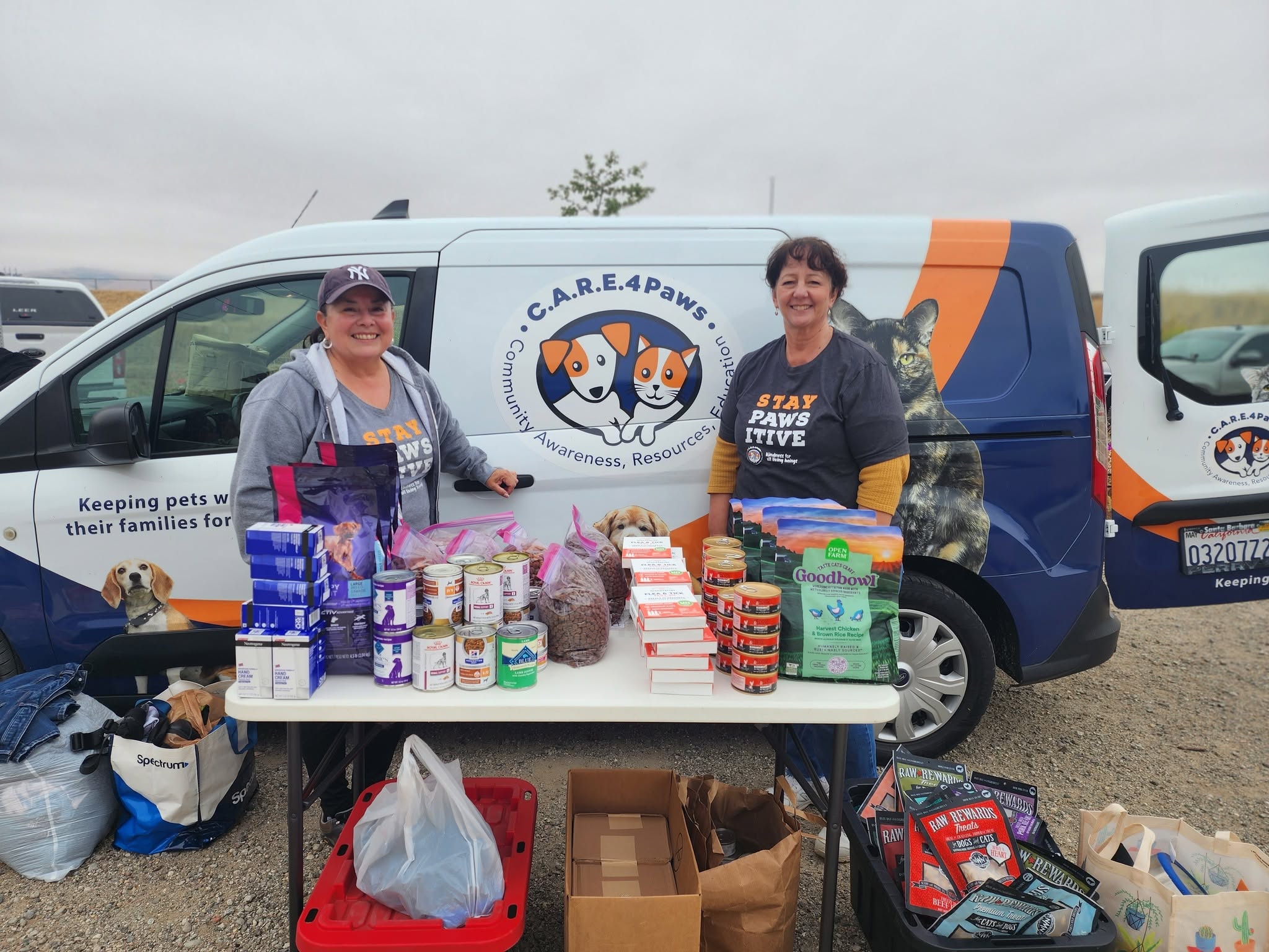 Two women stand behind a table with pet supplies, smiling, in front of a van with pet rescue logo, outdoors.