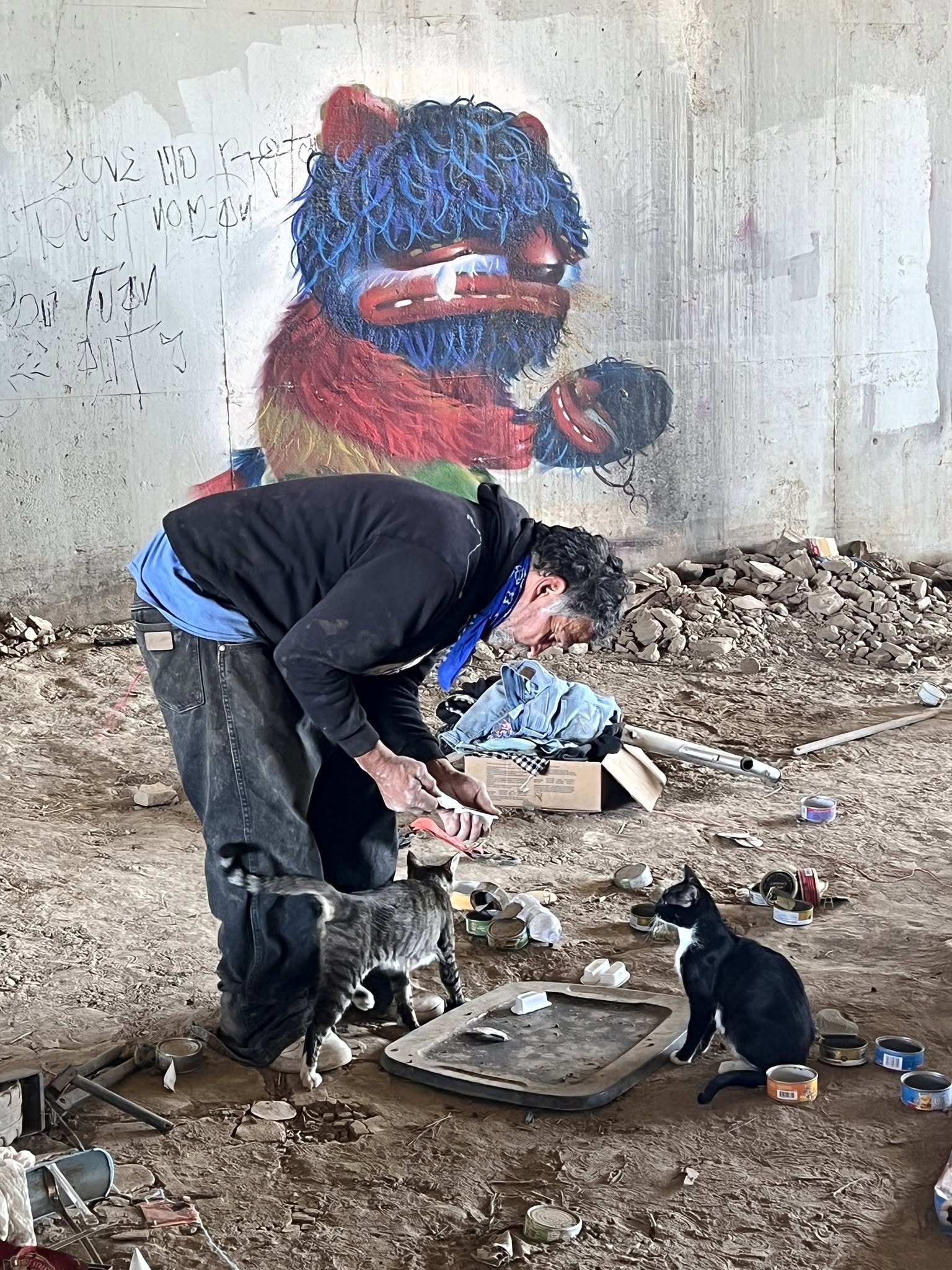 Man crouching on ground with two dogs, graffiti of a colorful creature on wall behind him, dirt and debris around.