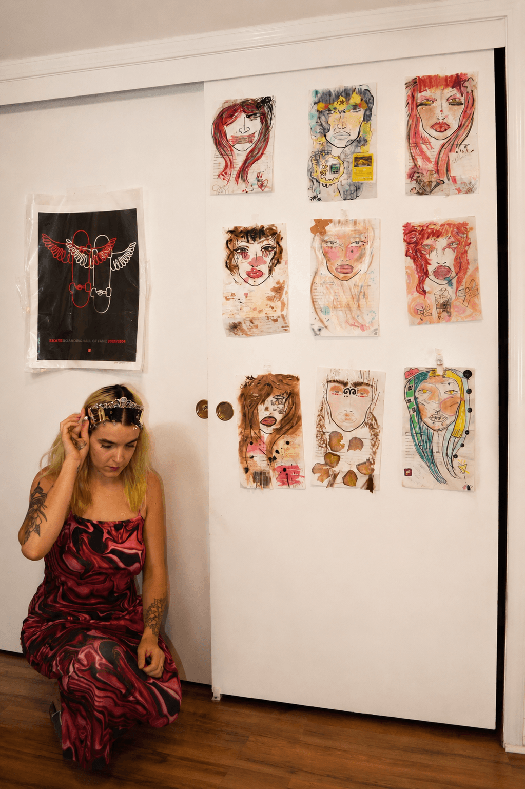 Young woman with long hair and a headband, wearing a red and black dress, kneeling in front of a white wall with colorful artwork.