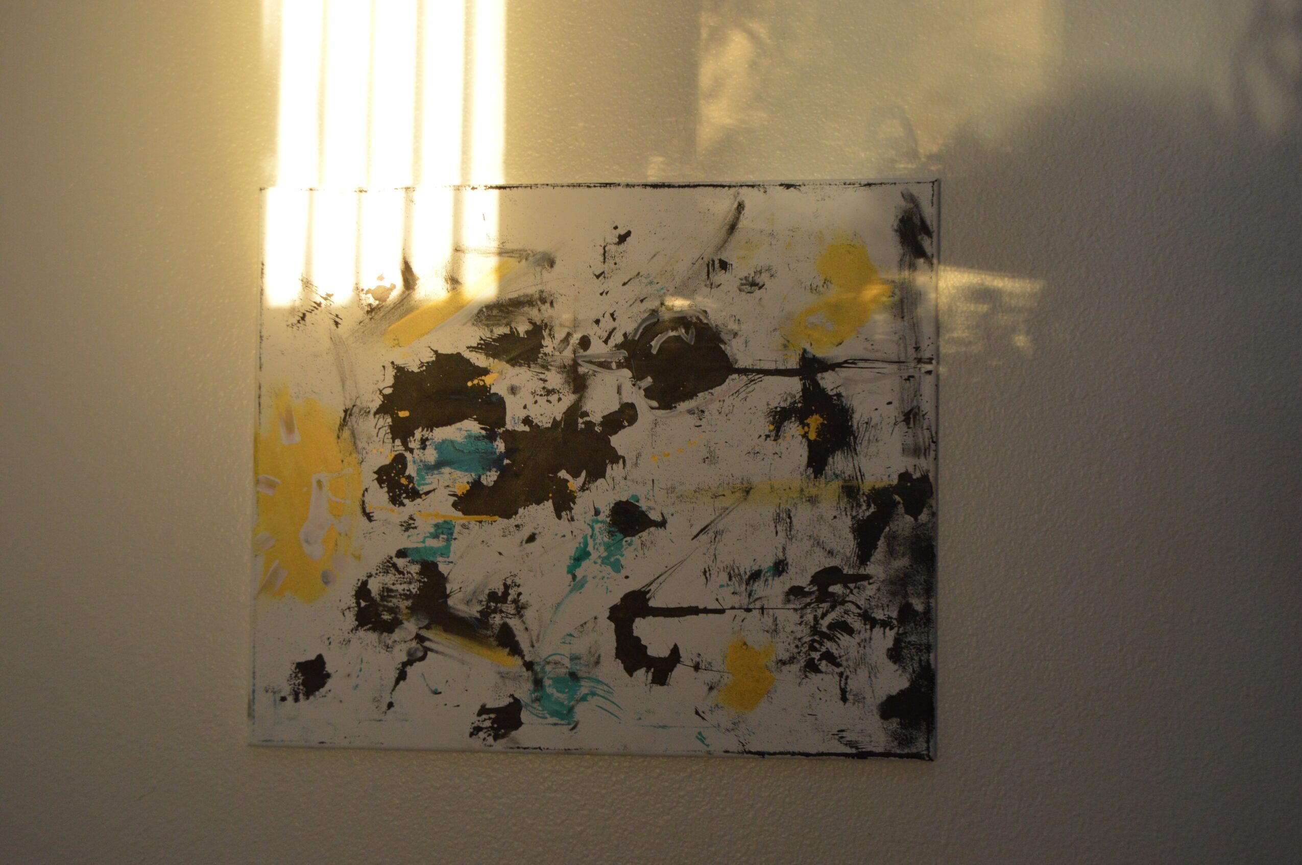 Abstract painting with black, yellow, and teal splashes on a white background, hung on a wall with light reflection.