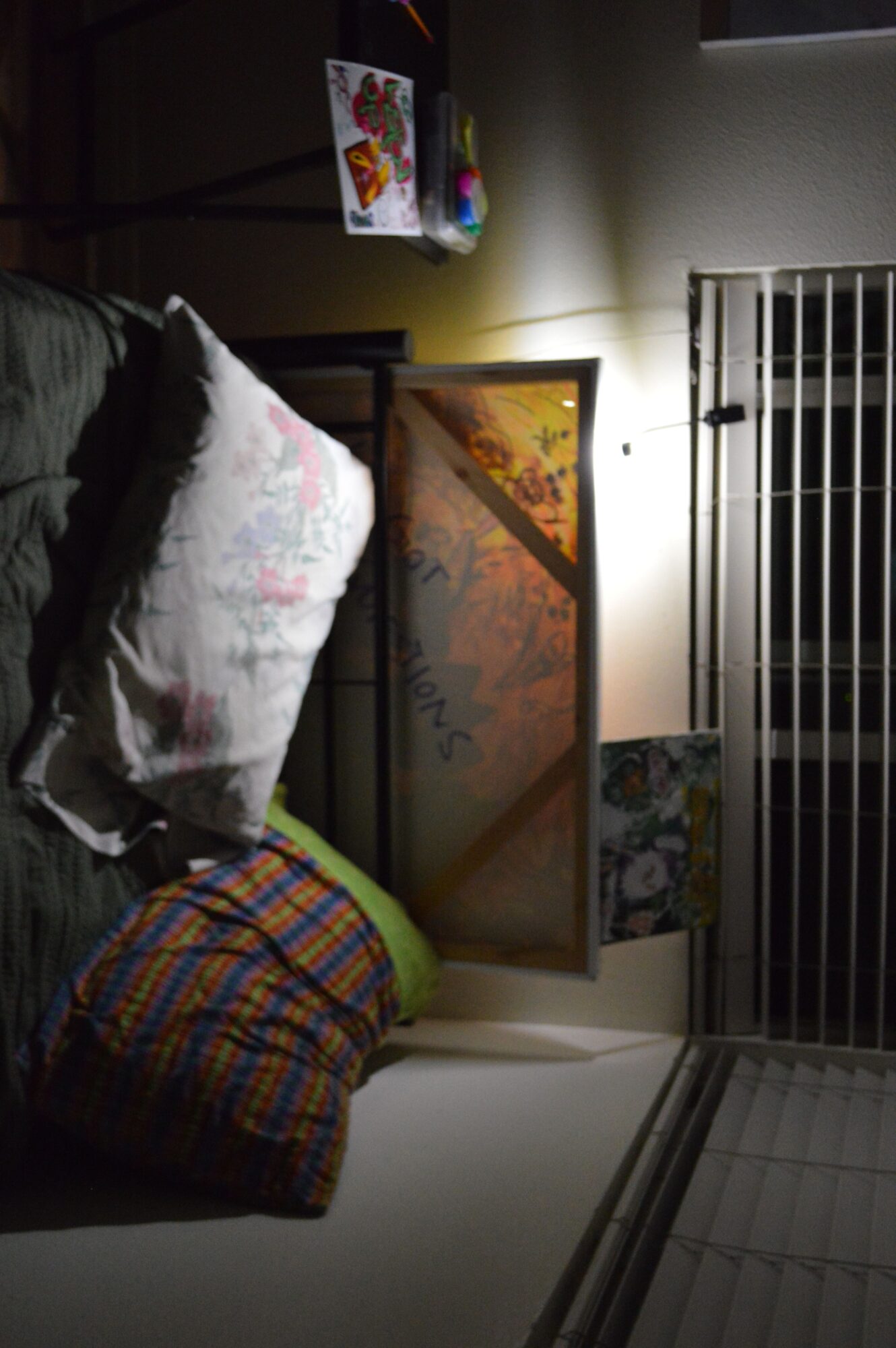 A bed with a pillow and a colorful blanket, a small table with items, and a window with blinds in a dimly lit room.