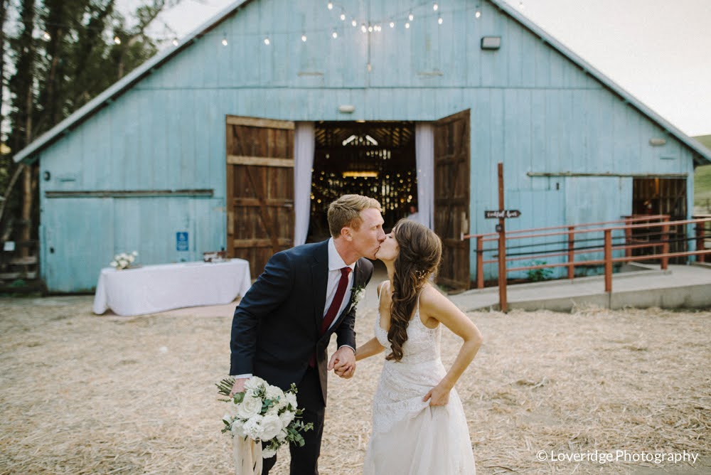 A bride and groom kiss outdoors in front of a blue barn, holding hands, with a table nearby, during a wedding.