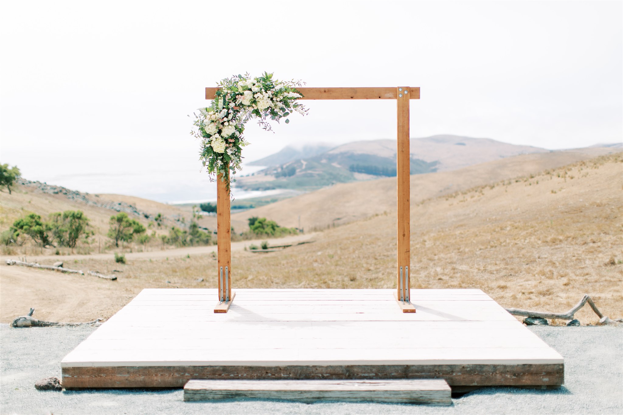 Empty outdoor stage with wooden arch decorated with flowers, set in a hilly landscape, overcast sky, white platform, no people present.
