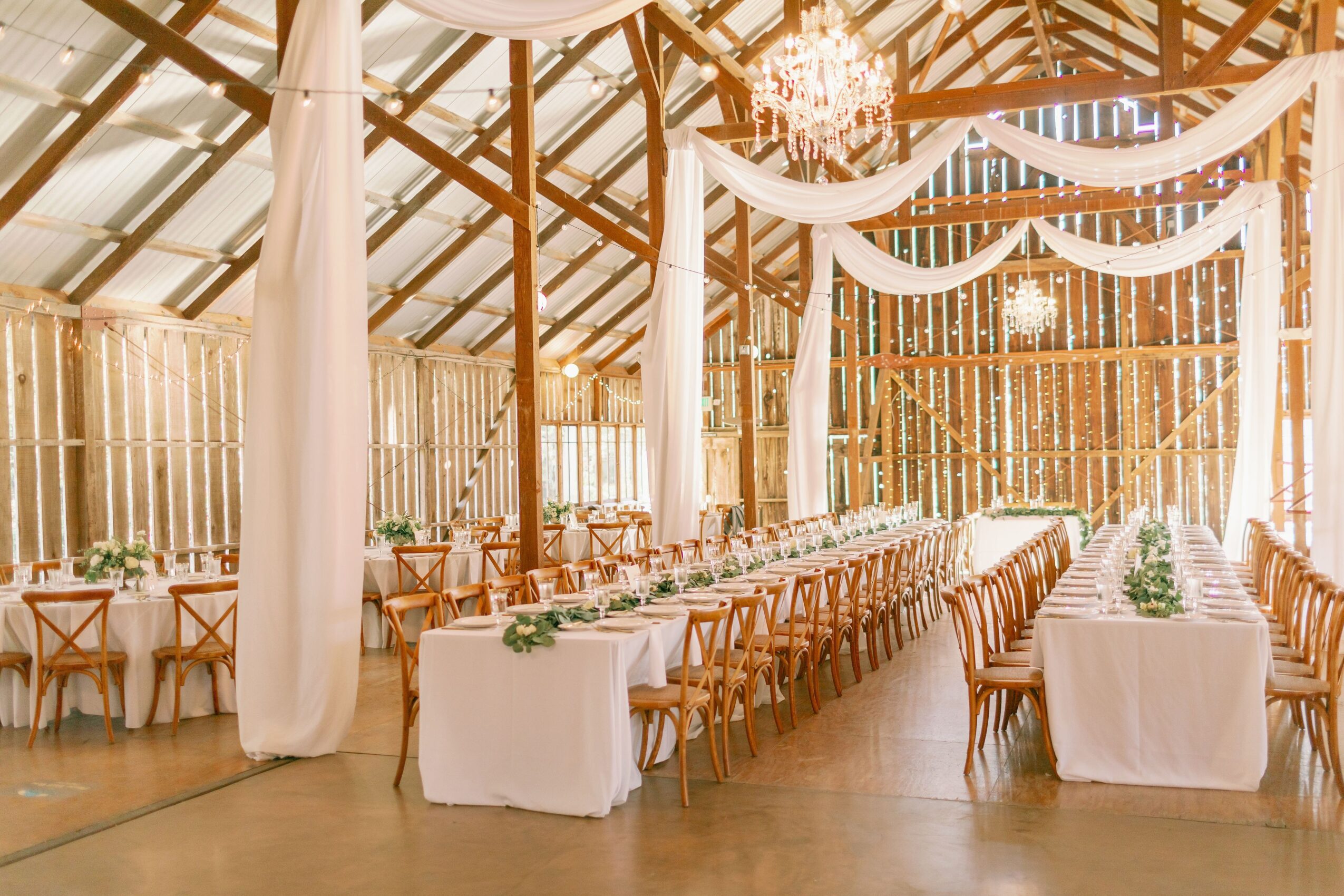 Elegant event space with long tables, chairs, draped fabric, chandeliers, and wooden walls, set for a celebration.