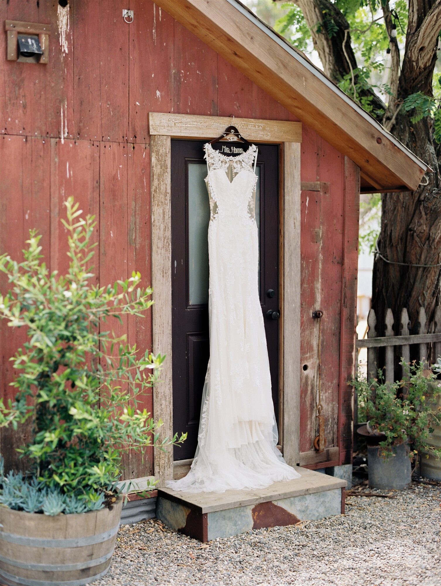 White wedding dress hanging on a door outside a rustic red wooden building, surrounded by plants and trees.