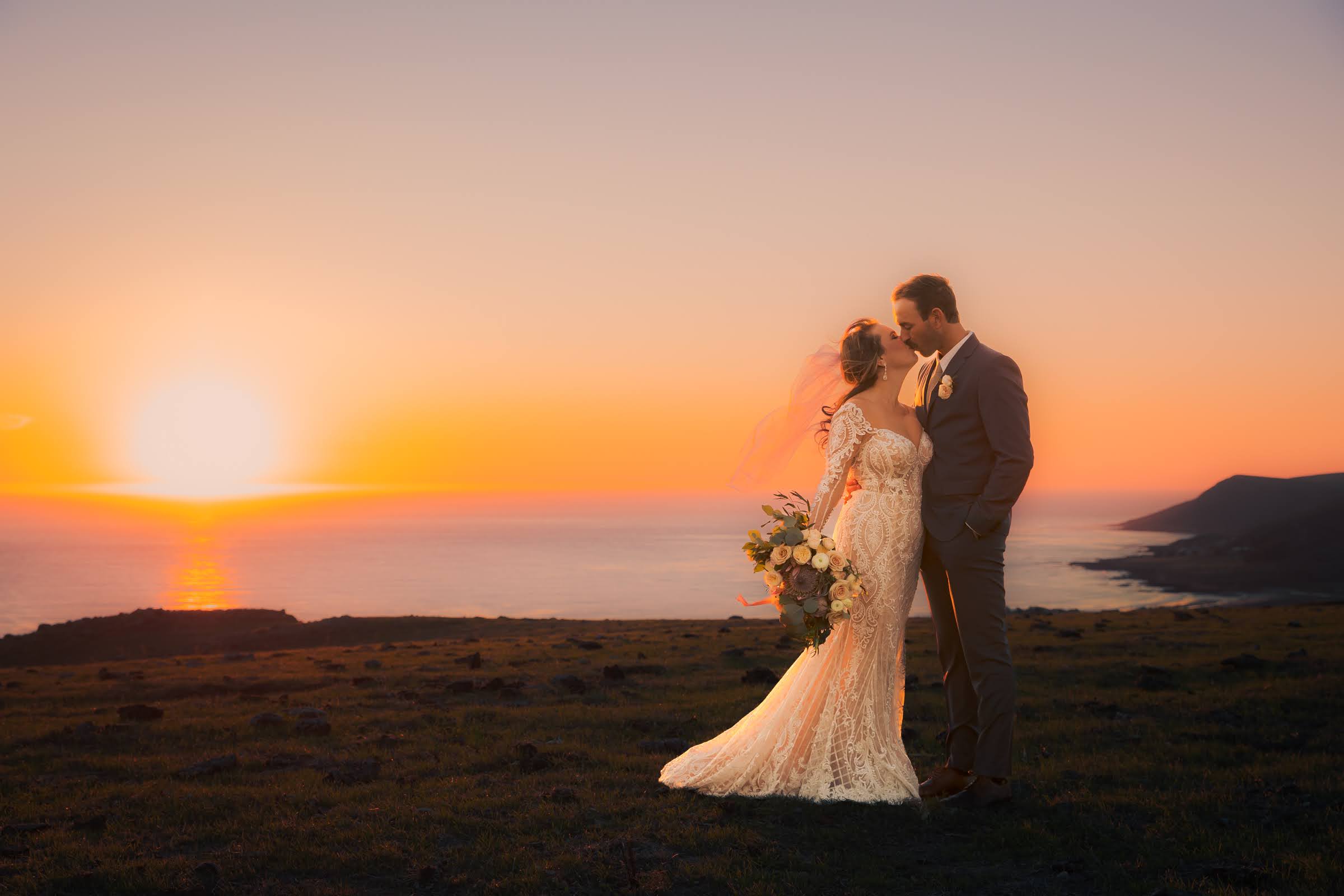 A bride and groom stand close together on a grassy area during sunset, with a body of water and hills in the background.