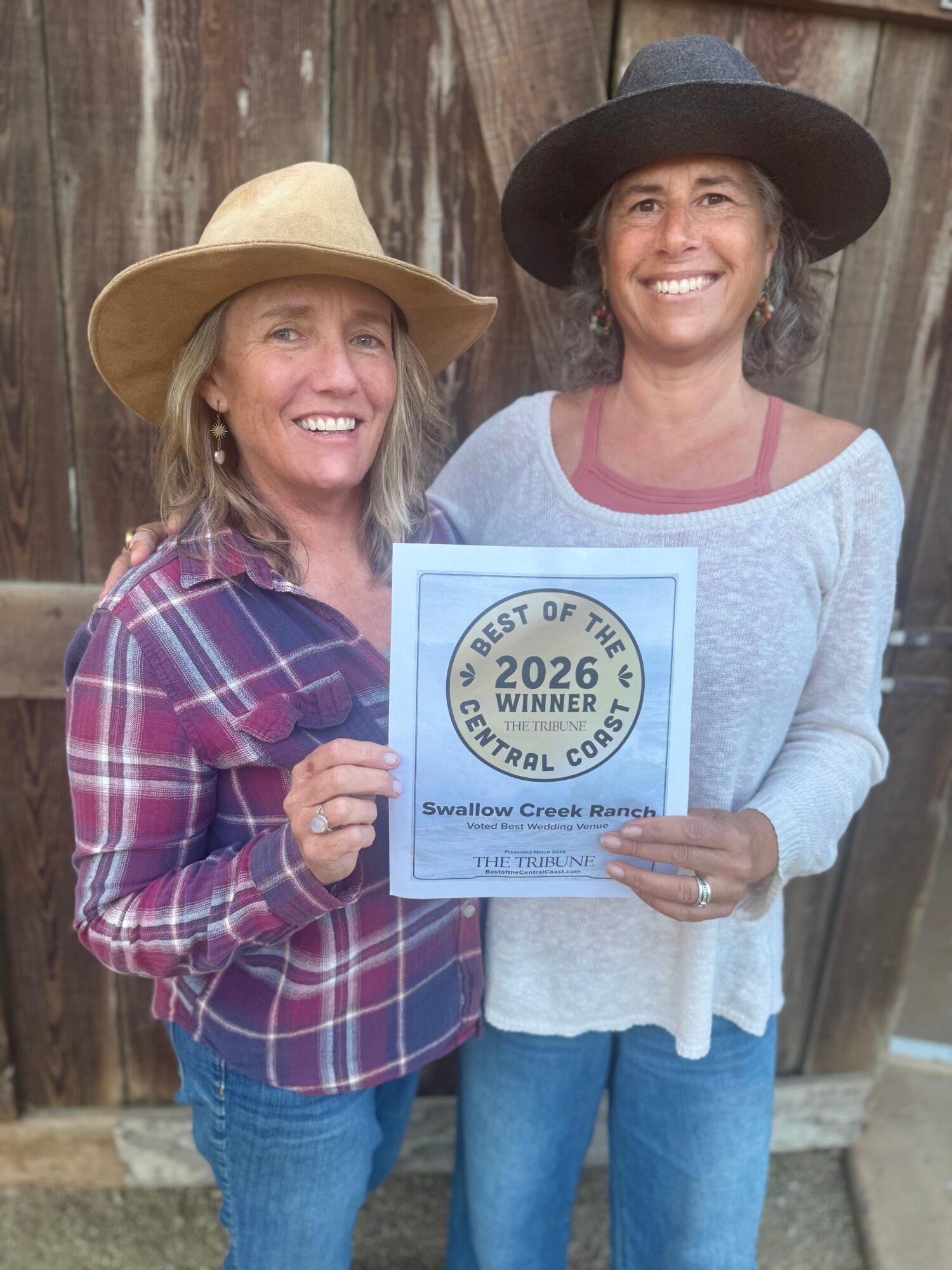 Two women smiling, holding a certificate, standing outdoors in front of a wooden fence, wearing hats and casual clothing.