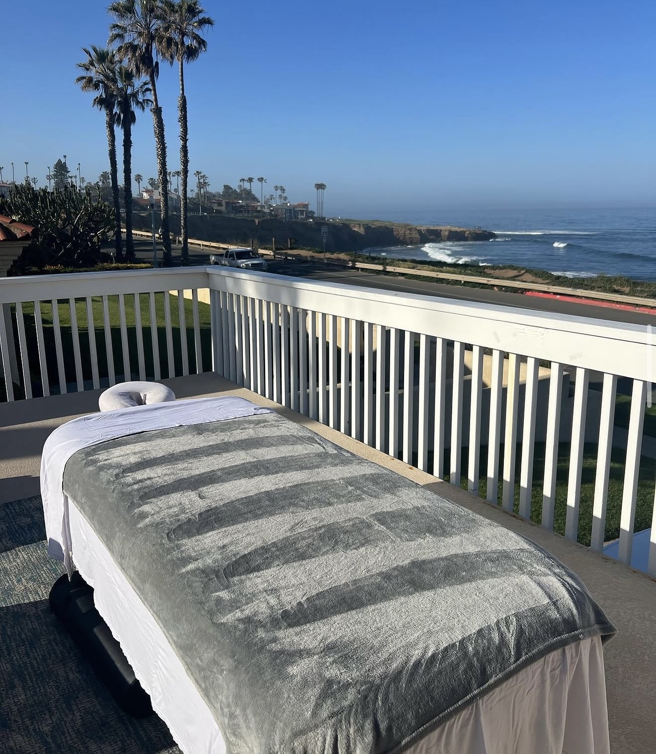 Massage table on balcony overlooking ocean, palm trees, and coastline under clear blue sky.