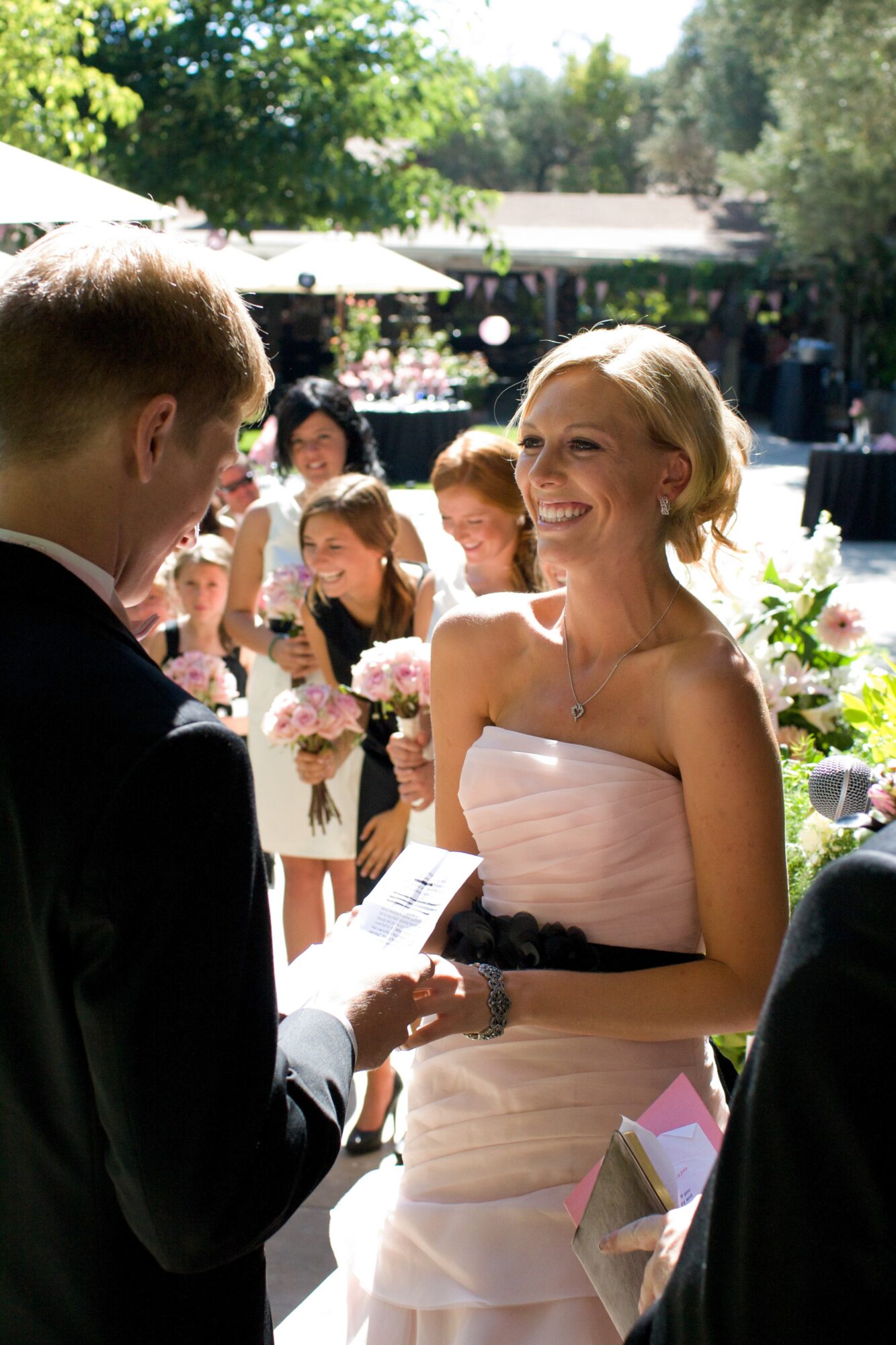 Woman in strapless dress smiling at man during outdoor wedding ceremony, guests in background holding flowers.