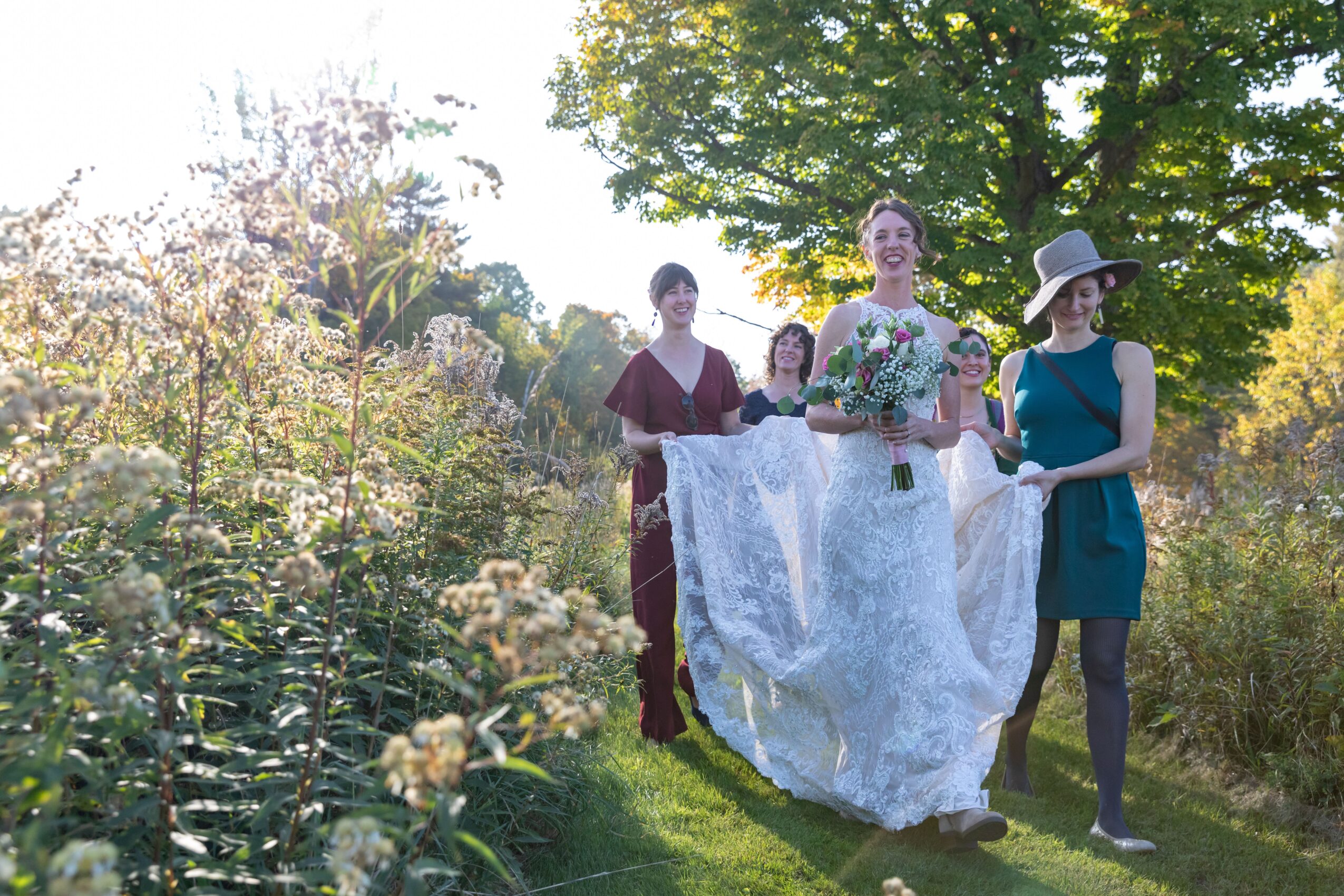 Four women walking outdoors on a sunny day, one in a wedding dress holding a bouquet, others in dresses and hat, surrounded by greenery.