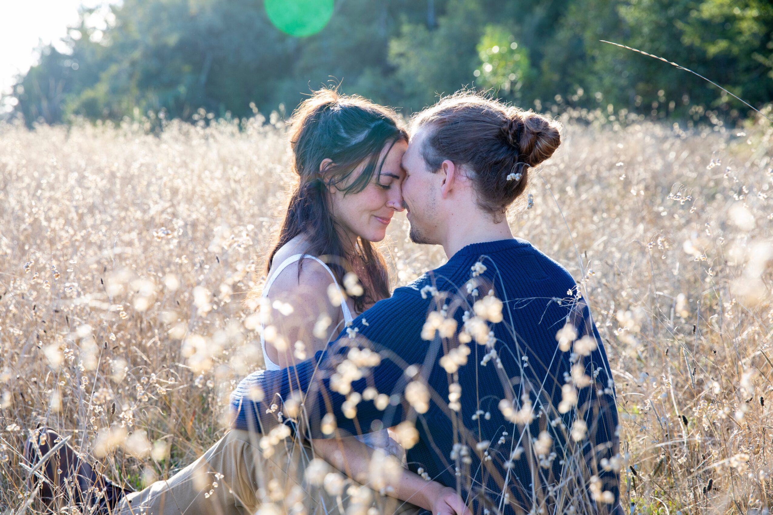 Two women face each other closely in a field of tall grass, with trees in the background, sunlight shining through.