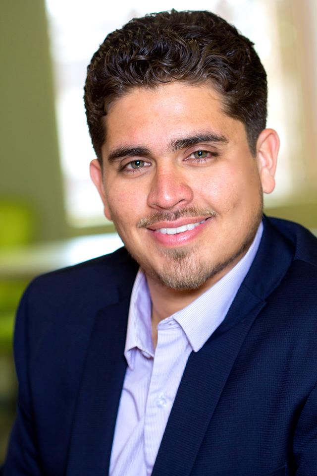 Young man with dark curly hair, light skin, wearing a navy blazer and white shirt, smiling, indoor background.