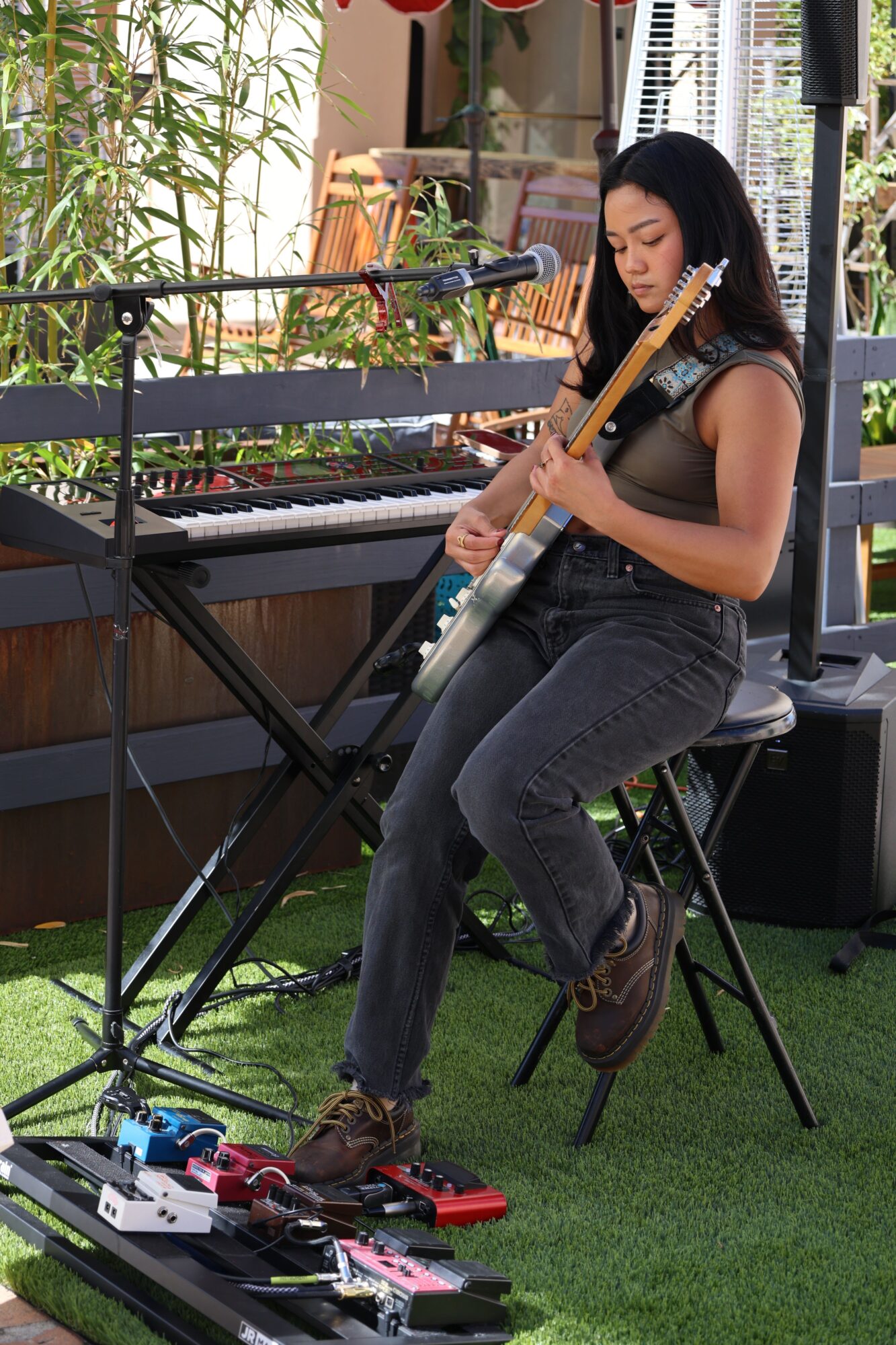 Woman sitting on a stool holding a guitar, surrounded by musical equipment and plants, in an outdoor setting.