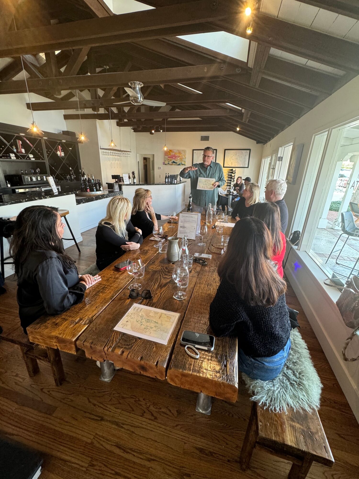 People gathered around a wooden table in a restaurant, with a woman standing and others seated, glasses and menus on the table.