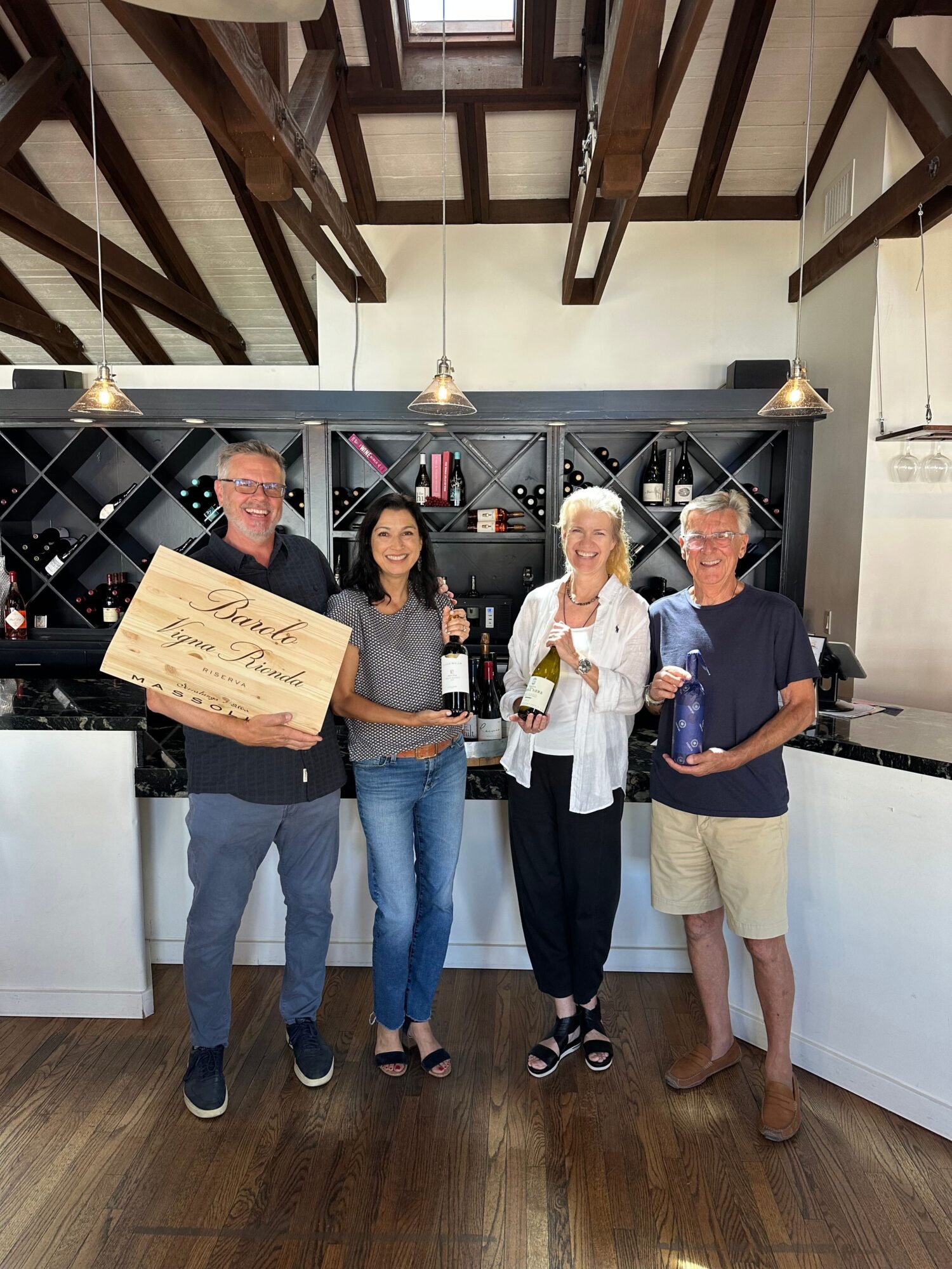 Four people standing in front of a wine rack, one holding a large check, in a room with wooden floors and ceiling beams.