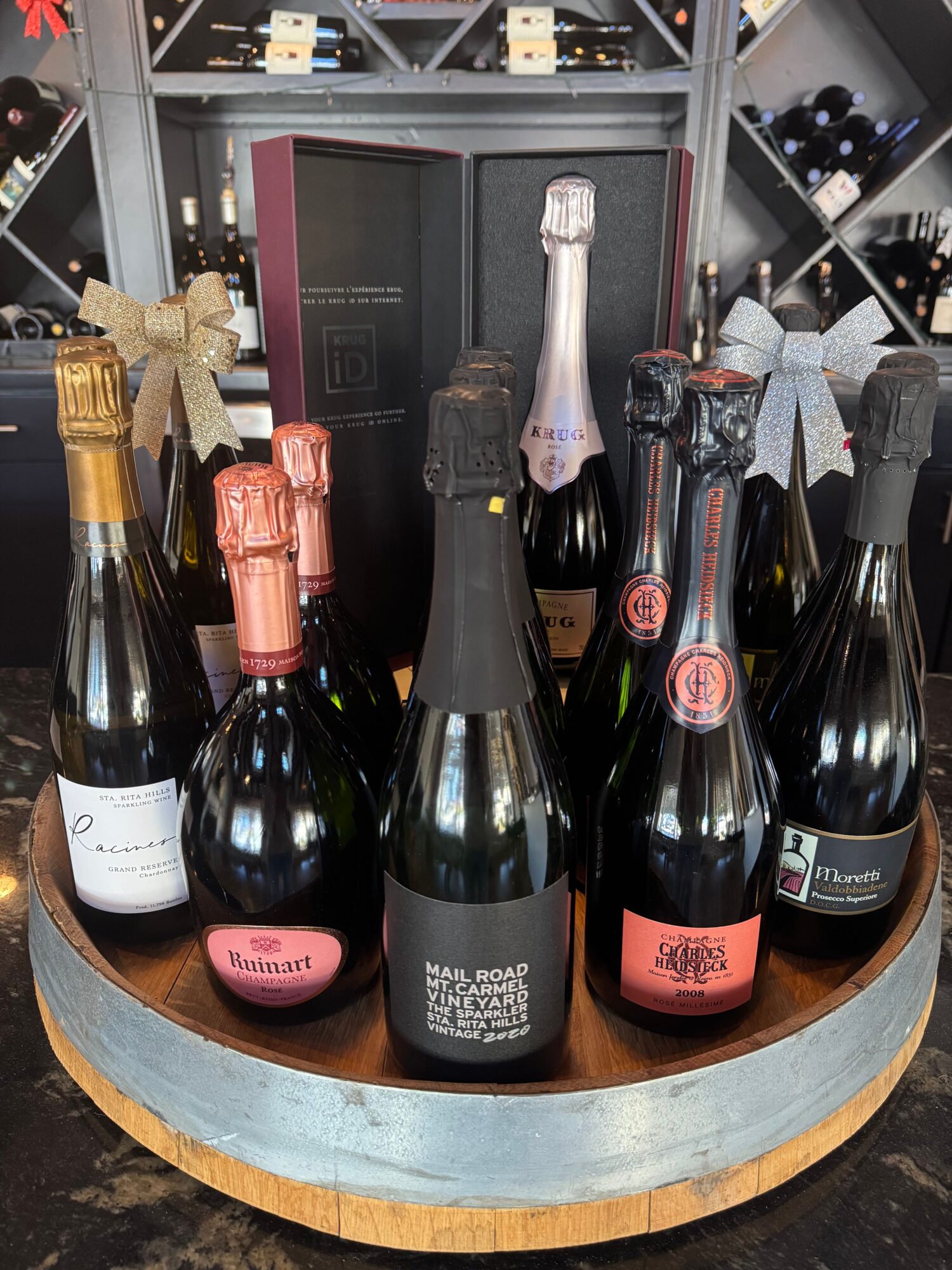 Six bottles of wine and champagne on a round wooden table, with a wine rack in the background.
