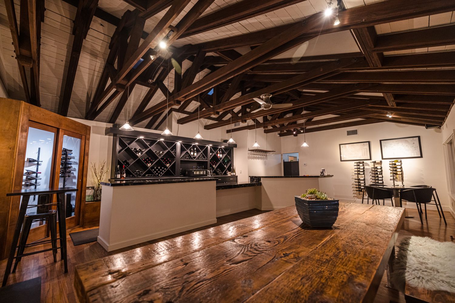 Interior of a bar with wooden ceiling beams, a counter with wine bottles, and seating area with tables and chairs.