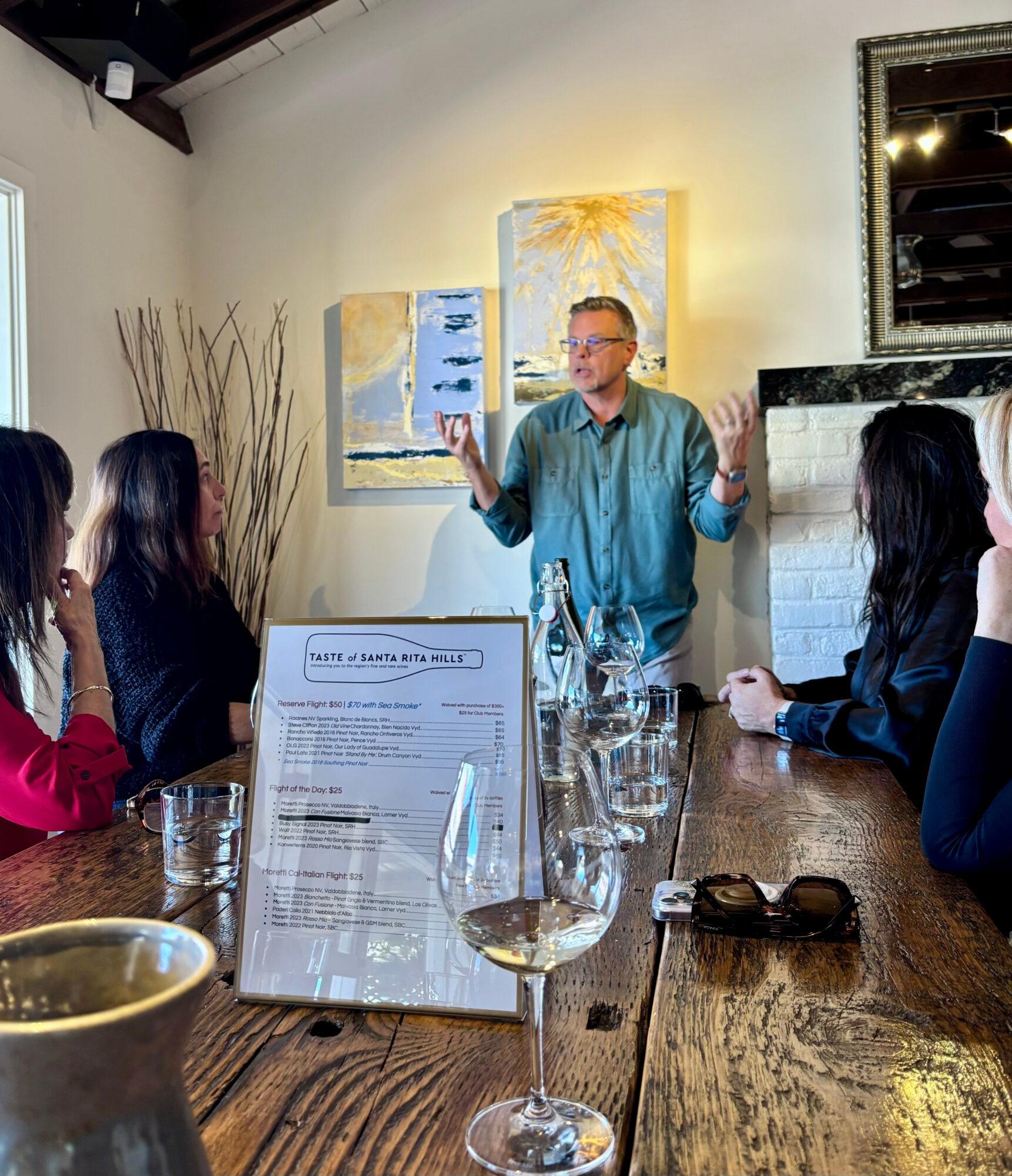Man speaking to a group of women seated around a wooden table in a room with artwork and a fireplace.
