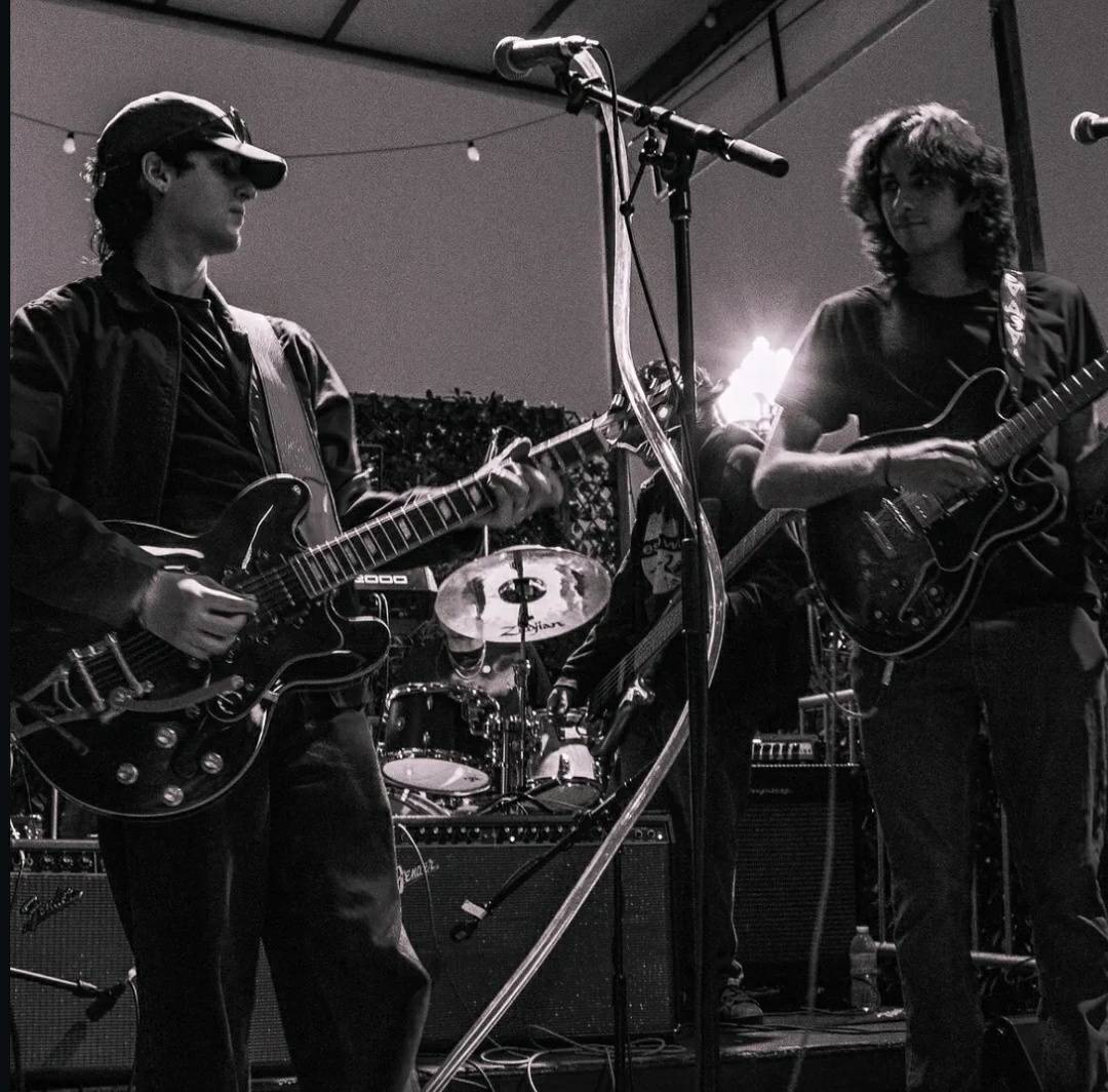 Two musicians playing guitars on stage with drums in background, stage lights shining behind them, black and white photo.