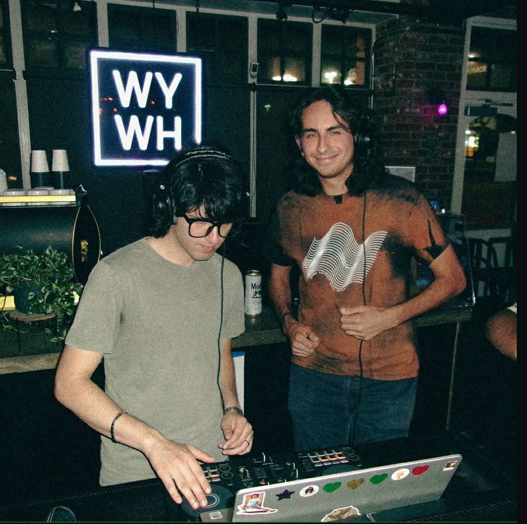 Two people, one DJ and one smiling person, in a dimly lit indoor setting with a neon sign in the background.