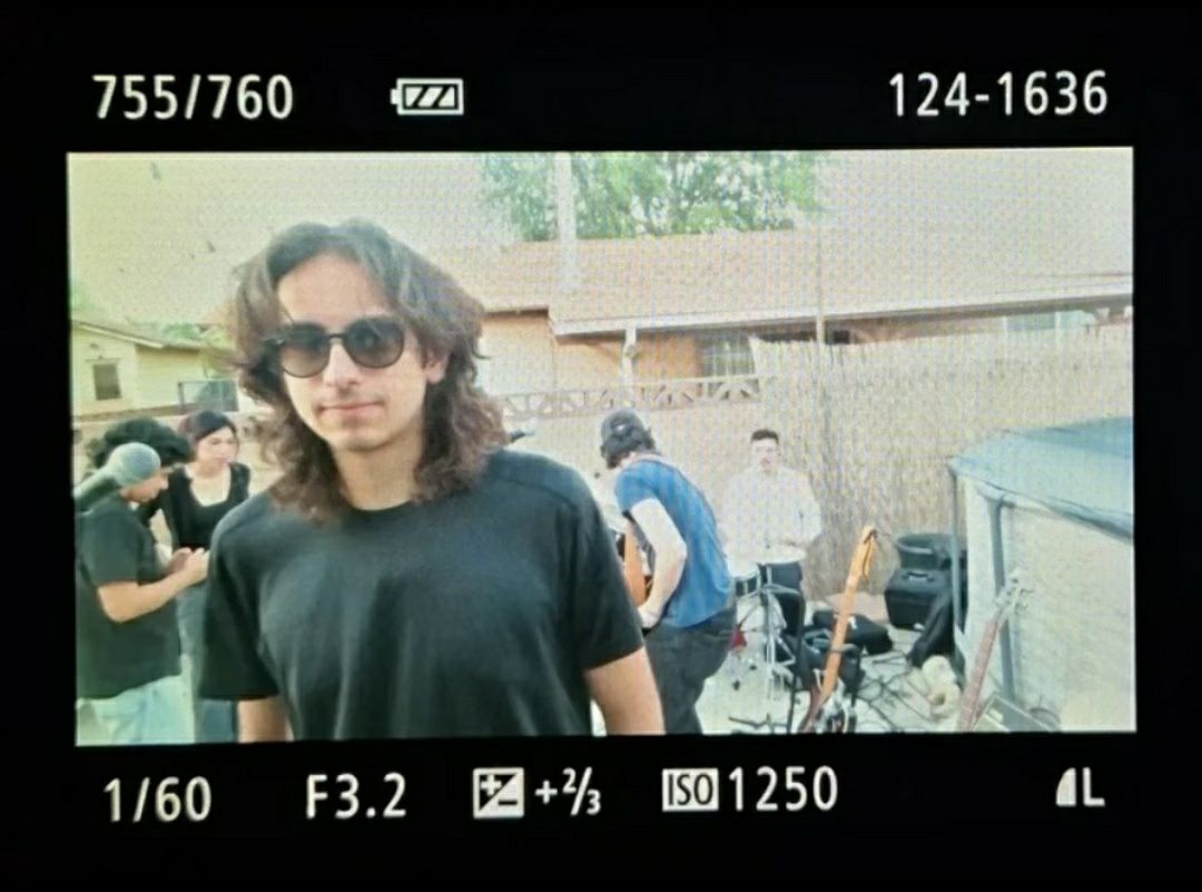 Person with curly hair and sunglasses outdoors, group of people and a fence in background.