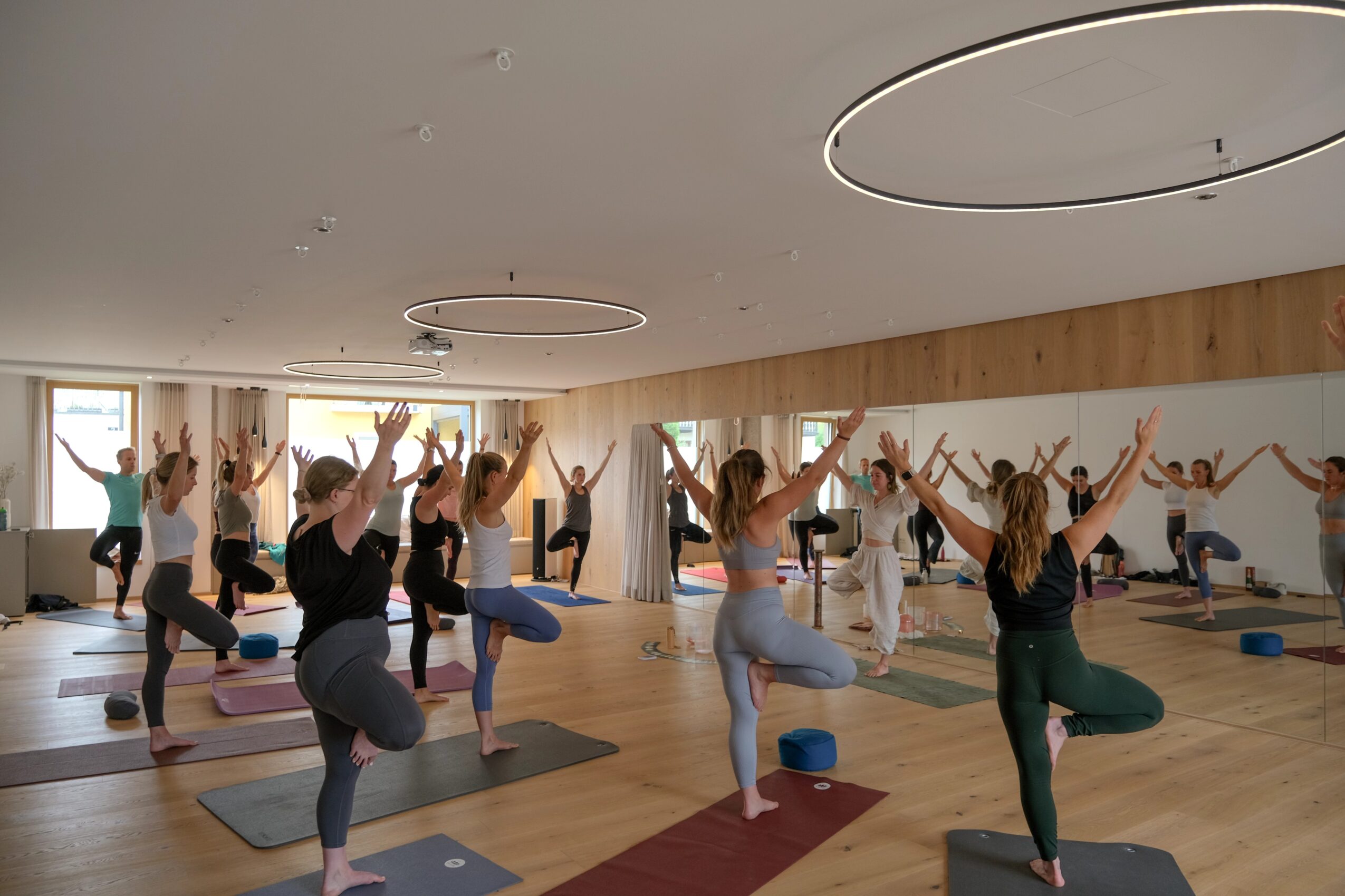 Group of people practicing yoga in a spacious, well-lit studio with wooden floors and large windows, some with arms raised.