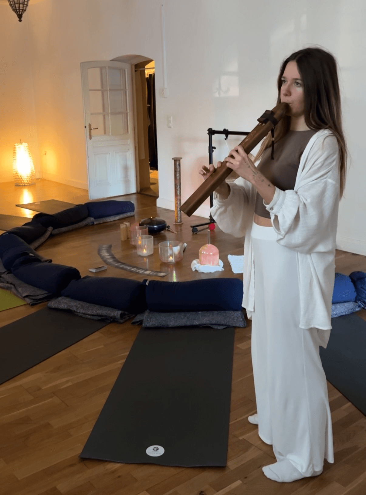 Young woman in white and gray clothing playing a wooden flute indoors, with yoga mats and a small table nearby.