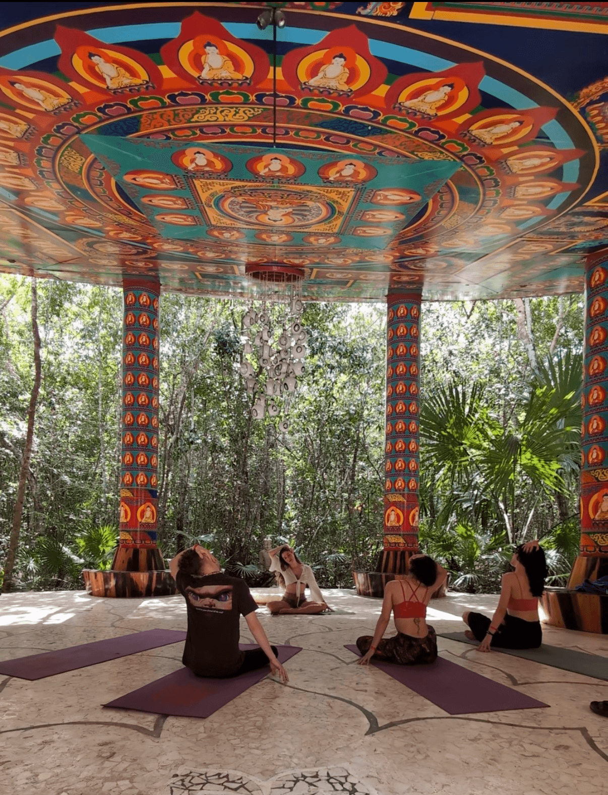 People practicing yoga on mats under a colorful, decorated canopy with trees in the background.