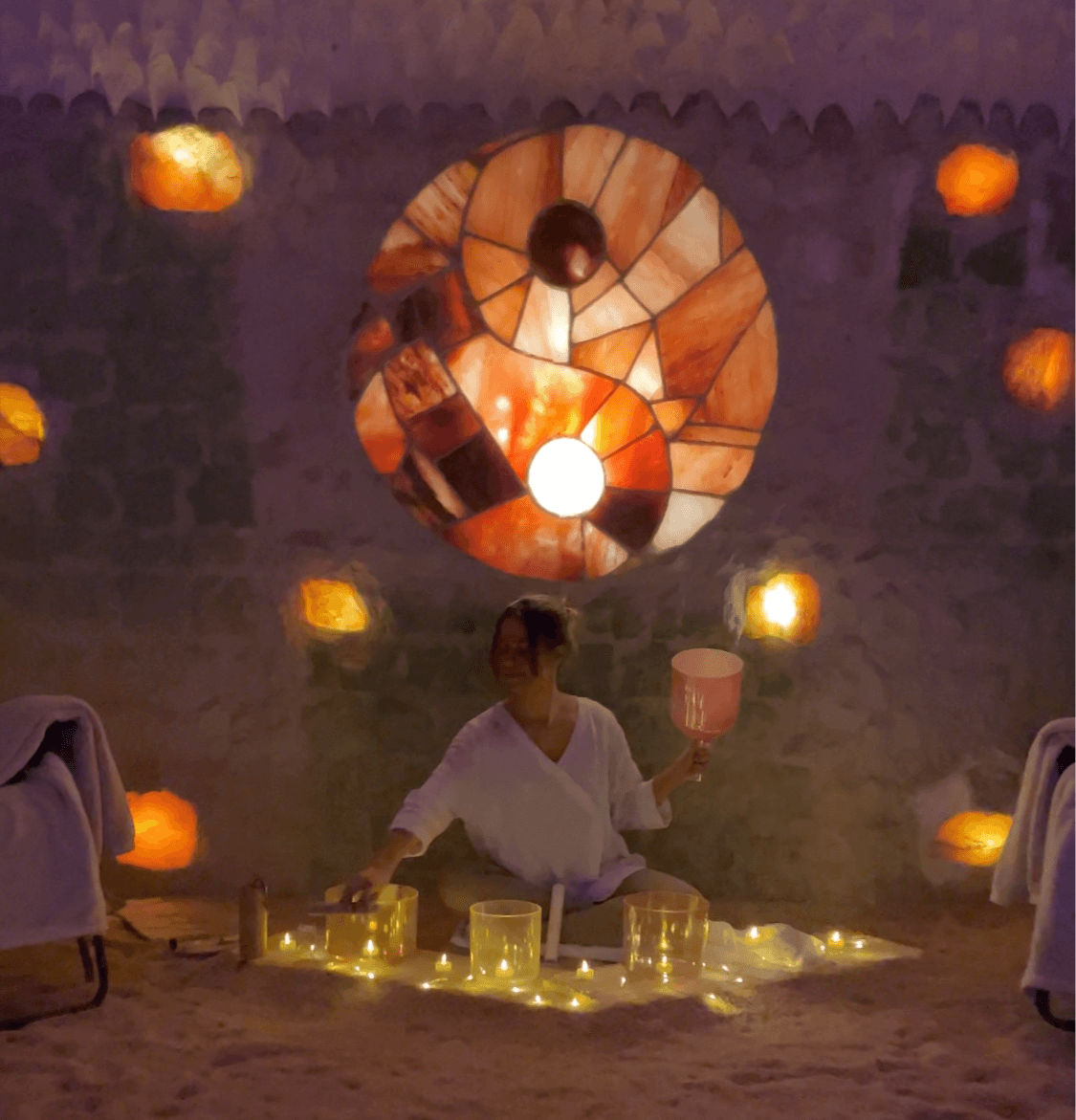 Person sitting cross-legged with a drum, surrounded by glowing cups, in a dimly lit room with a circular stained glass window.