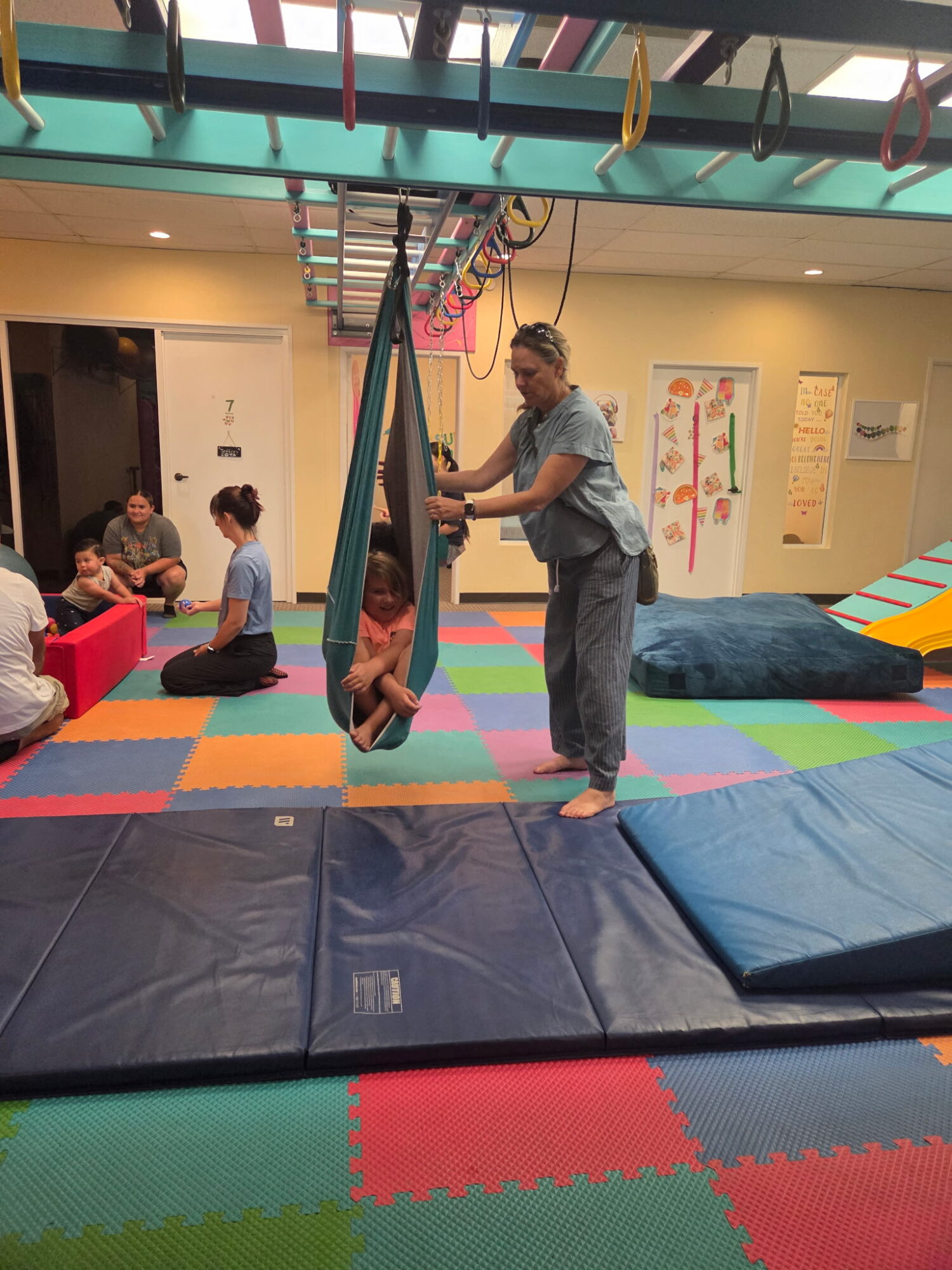 Child swinging on a hoop swing with adult supervising indoors, colorful mats on floor, other children in background.