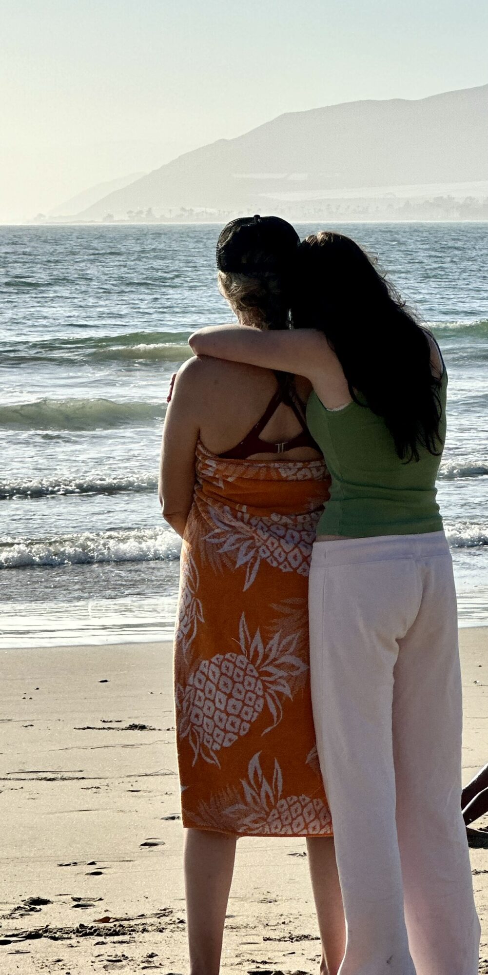 Two people hugging on a beach with ocean waves and mountains in the background.