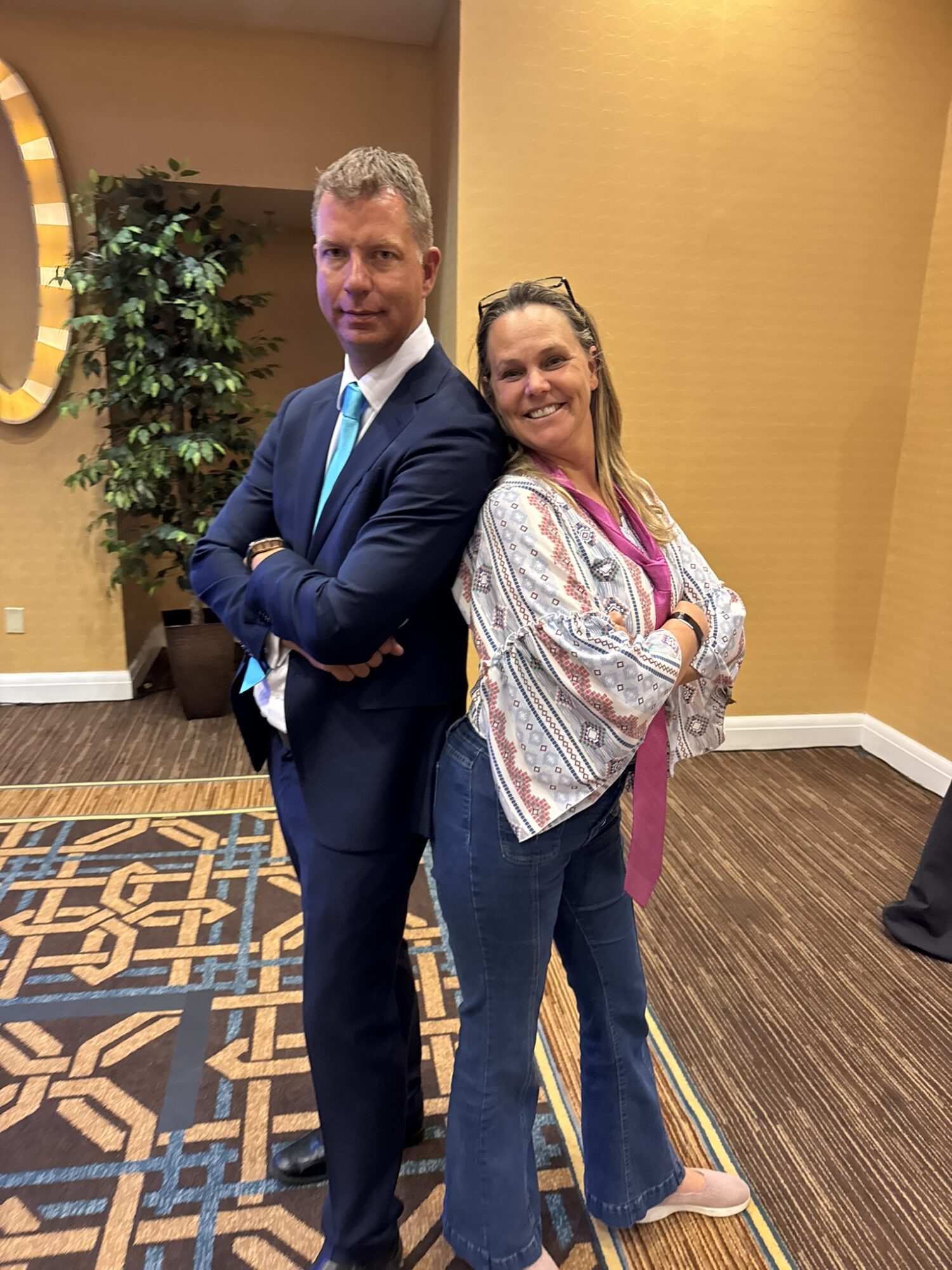 A man in a suit and a woman in casual clothing stand back-to-back with arms crossed in an indoor setting.