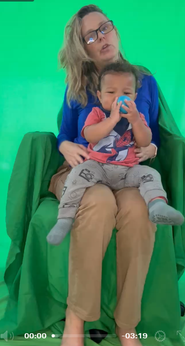 Woman with glasses holding a child on her lap, green screen background, woman wearing a blue top, child drinking from a cup.