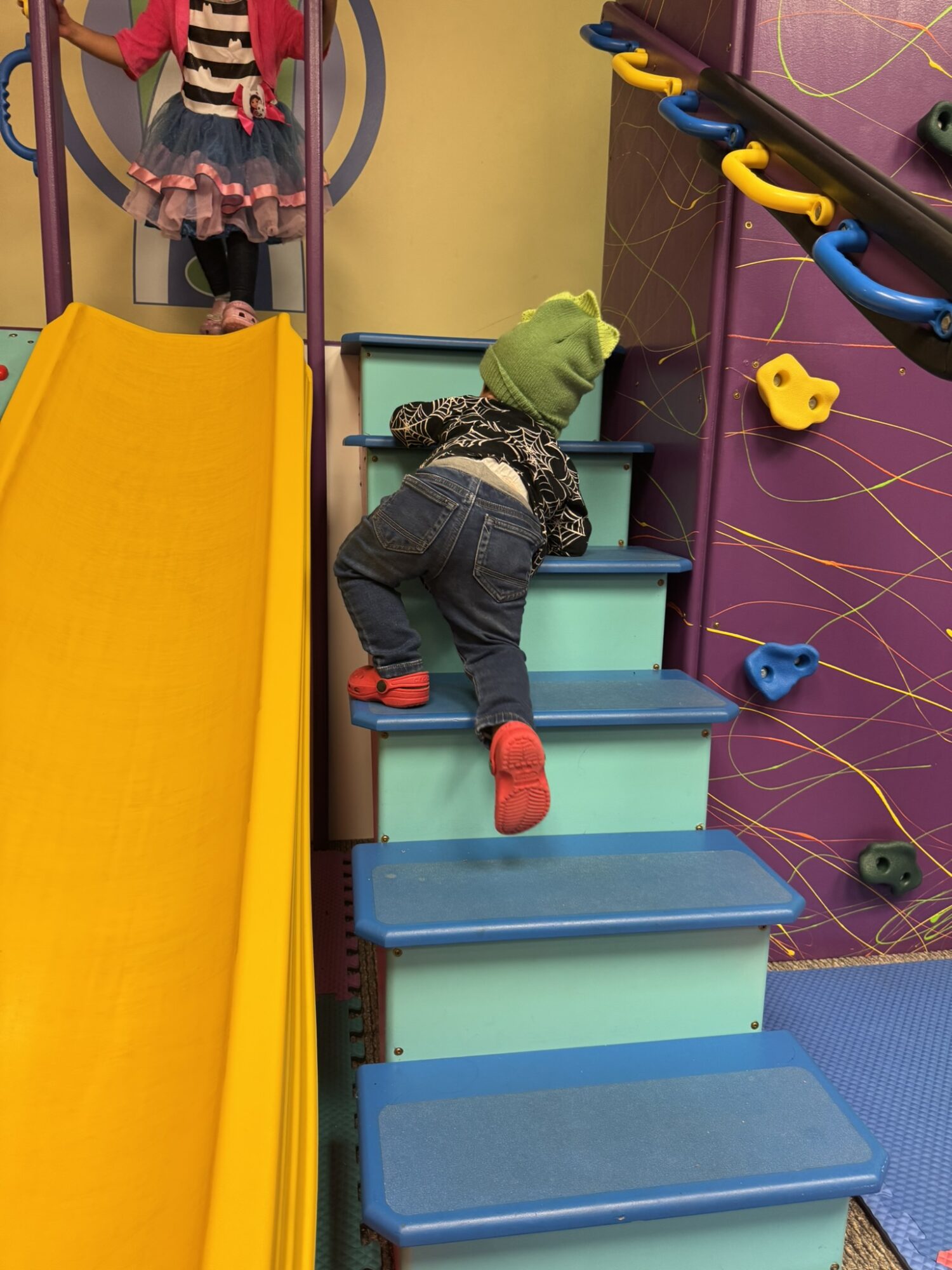 Child climbing stairs in an indoor play area, wearing a green hat, black and white shirt, jeans, and red shoes.