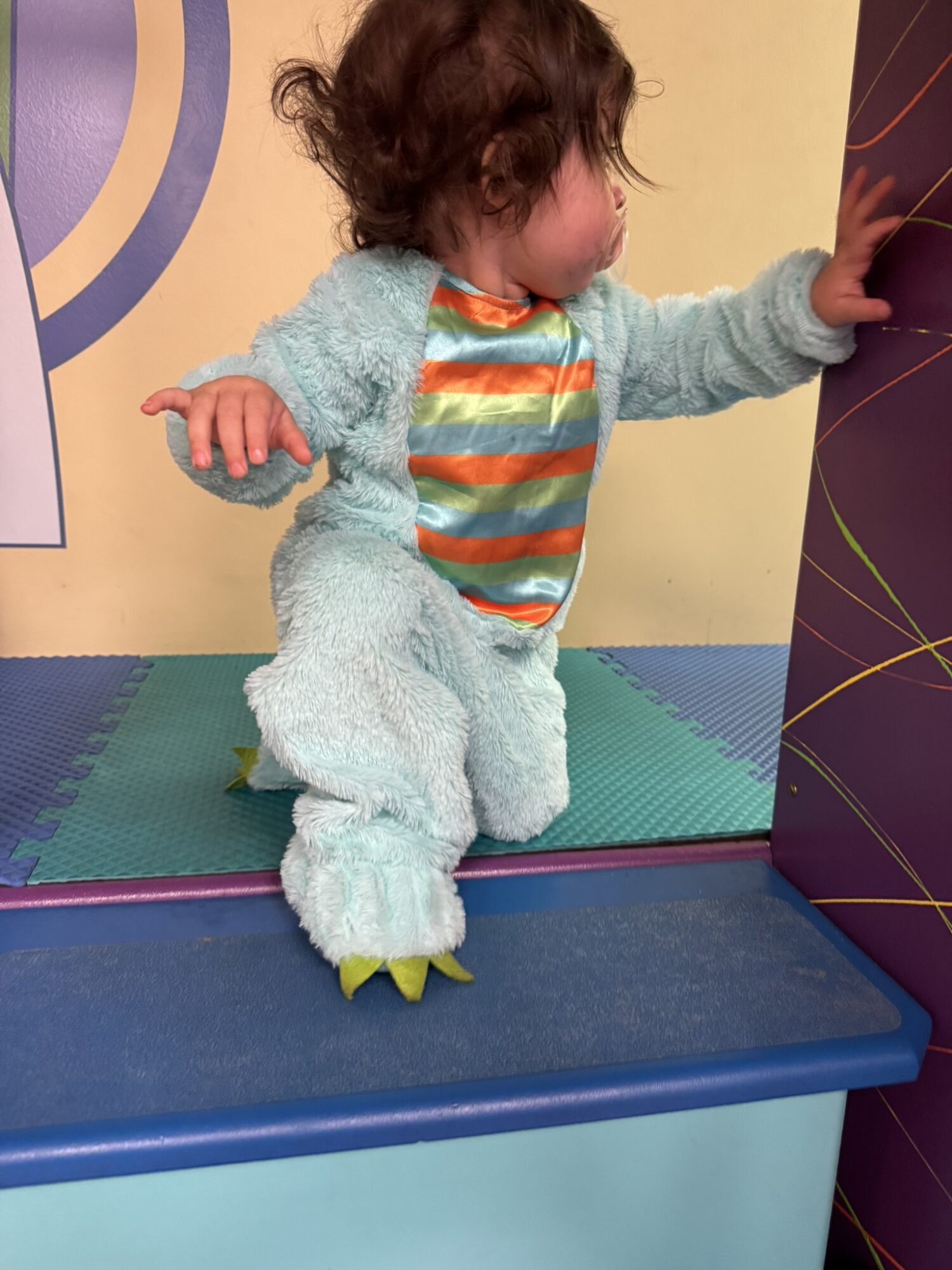 Child in a fluffy costume with yellow claws, standing on a blue surface, reaching out with one hand, facing sideways.