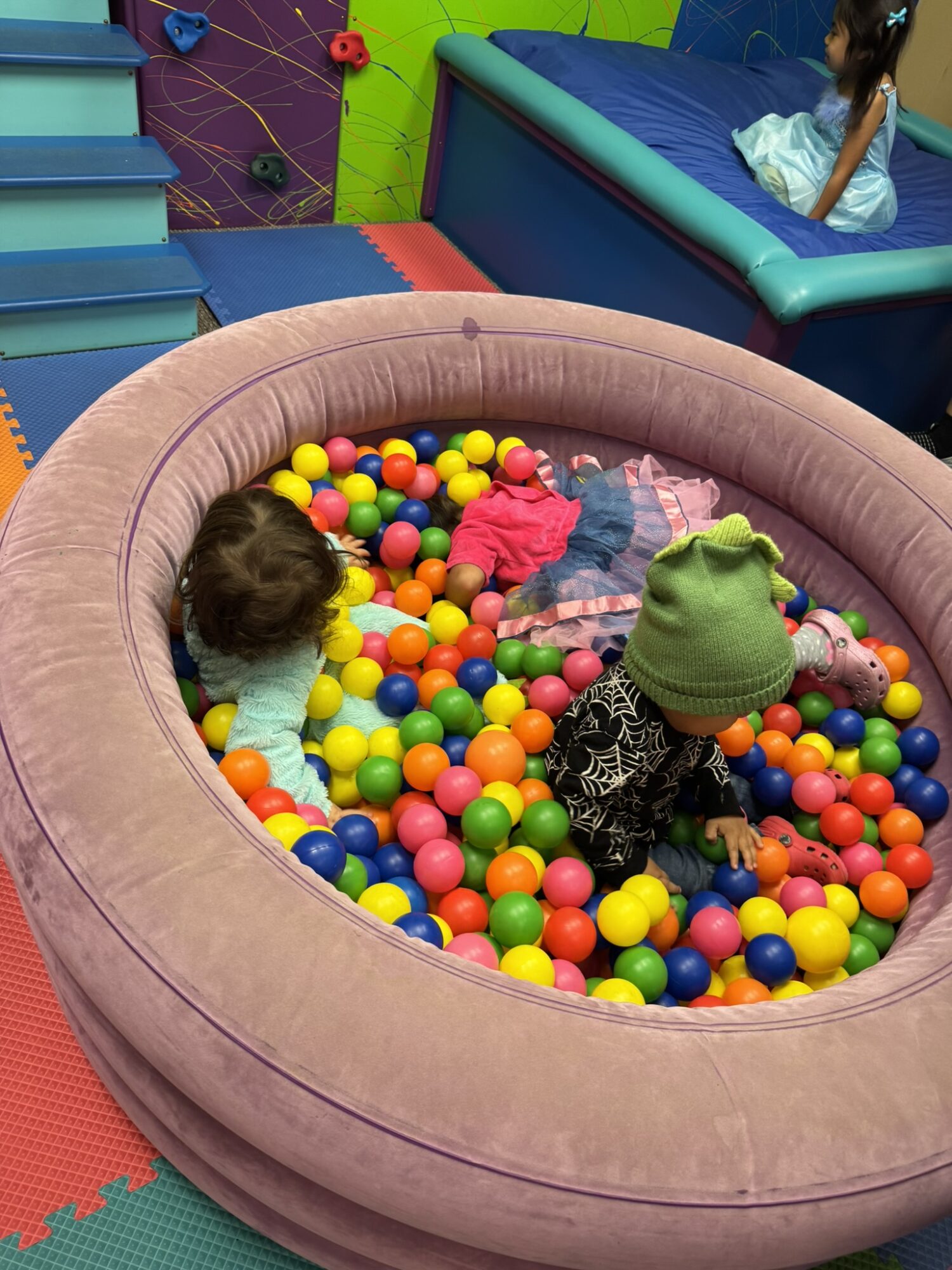 Two children playing in a round ball pit filled with colorful plastic balls, surrounded by soft padded walls.