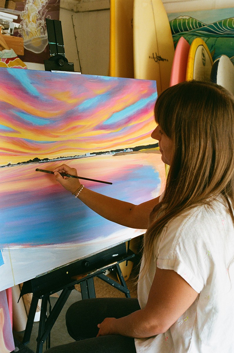 Woman painting a colorful sunset landscape on canvas, with surfboards and a yellow surfboard in the background.